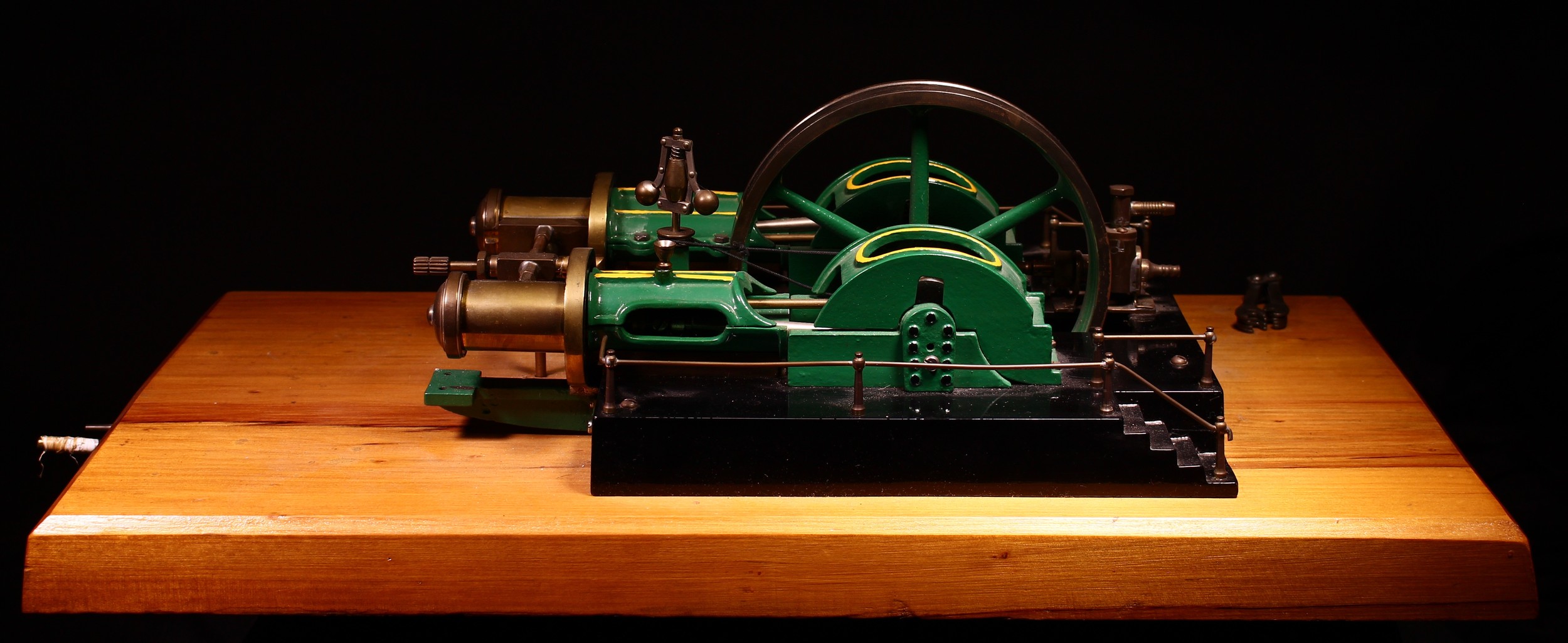 A well-engineered live steam horizontal stationary steam mill engine, central 6.5" diameter six spoke flywheel painted green, flanked by a pair of twin cylinder pistons, raised upon a black rectangular platform with a pair of staircases and handrails, mounted upon a rectangular shaped bespoke wooden plinth, 20cm high, 56.5cm wide, 29.5cm deep