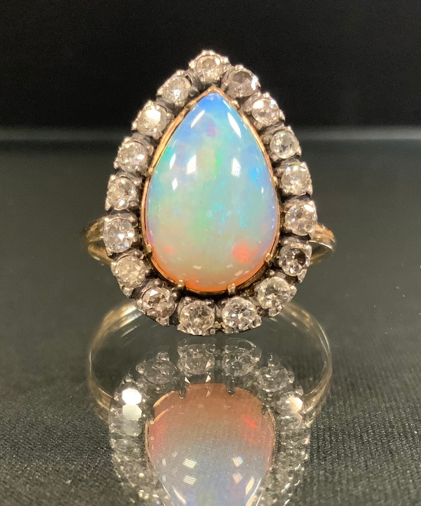 An opal and diamond ring, large pear cabochon opal, flashing vibrant green, blue and violet colour play, measuring approx 15mm x 9.5mm, surrounded by seventeen old brilliant cut diamonds, total estimated diamond weight approx 1,0ct, unmarked gold coloured metal shank, size R, 4.6g gross
