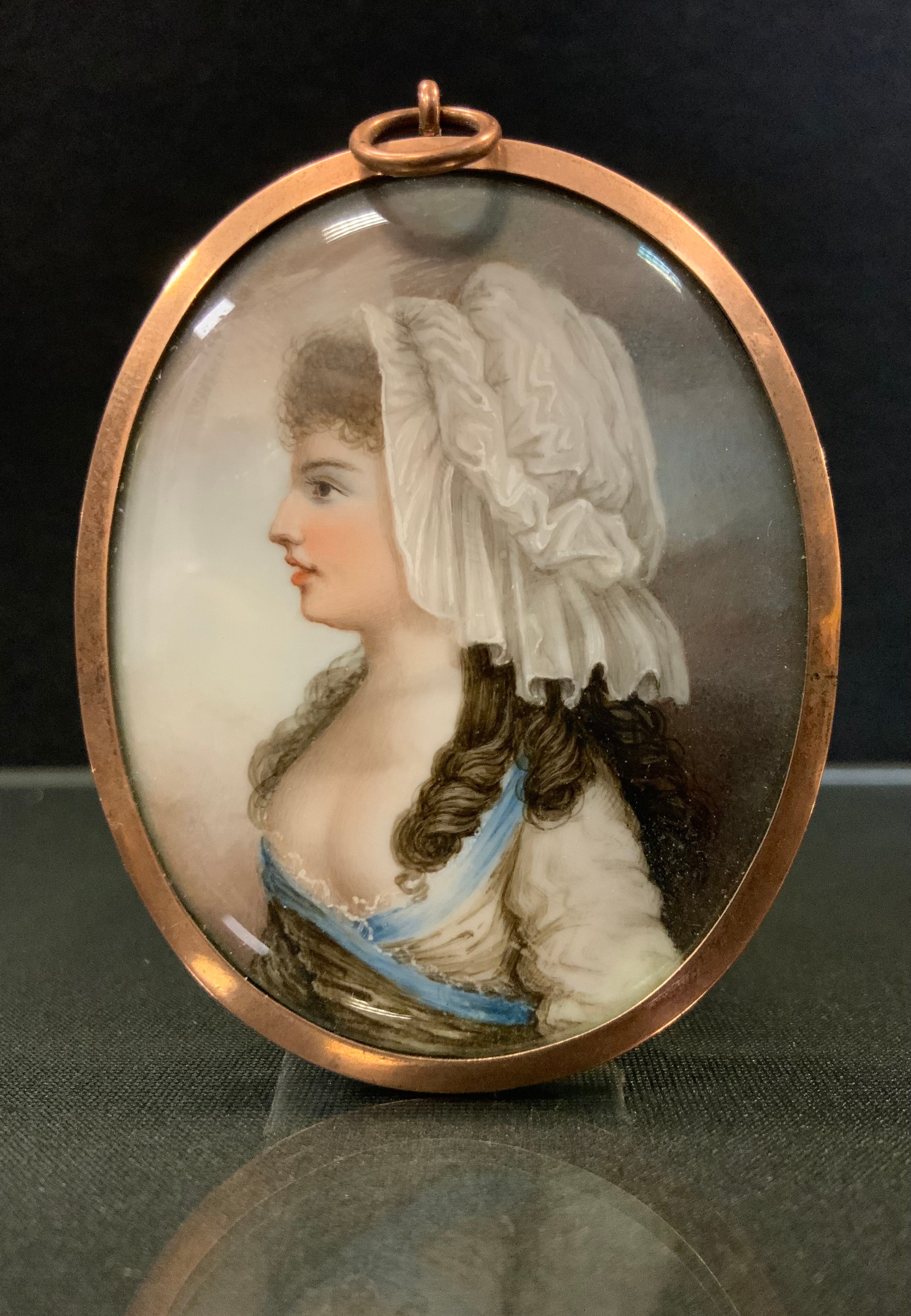 English School (19th century), a portrait miniature, of a lady, half length, wearing a cotton bonnet, blue strap and white dress,   watercolour on ivory, oval, 6cm x 4.5cm, gold plated frame, the verso in blue enamel  This item has been registered as exempt from the UK Ivory Act 2018, on account of it being a portrait miniature made before 1918 with a surface area less than 320 square centimetres.   Ivory declaration submission reference: UYF959QM