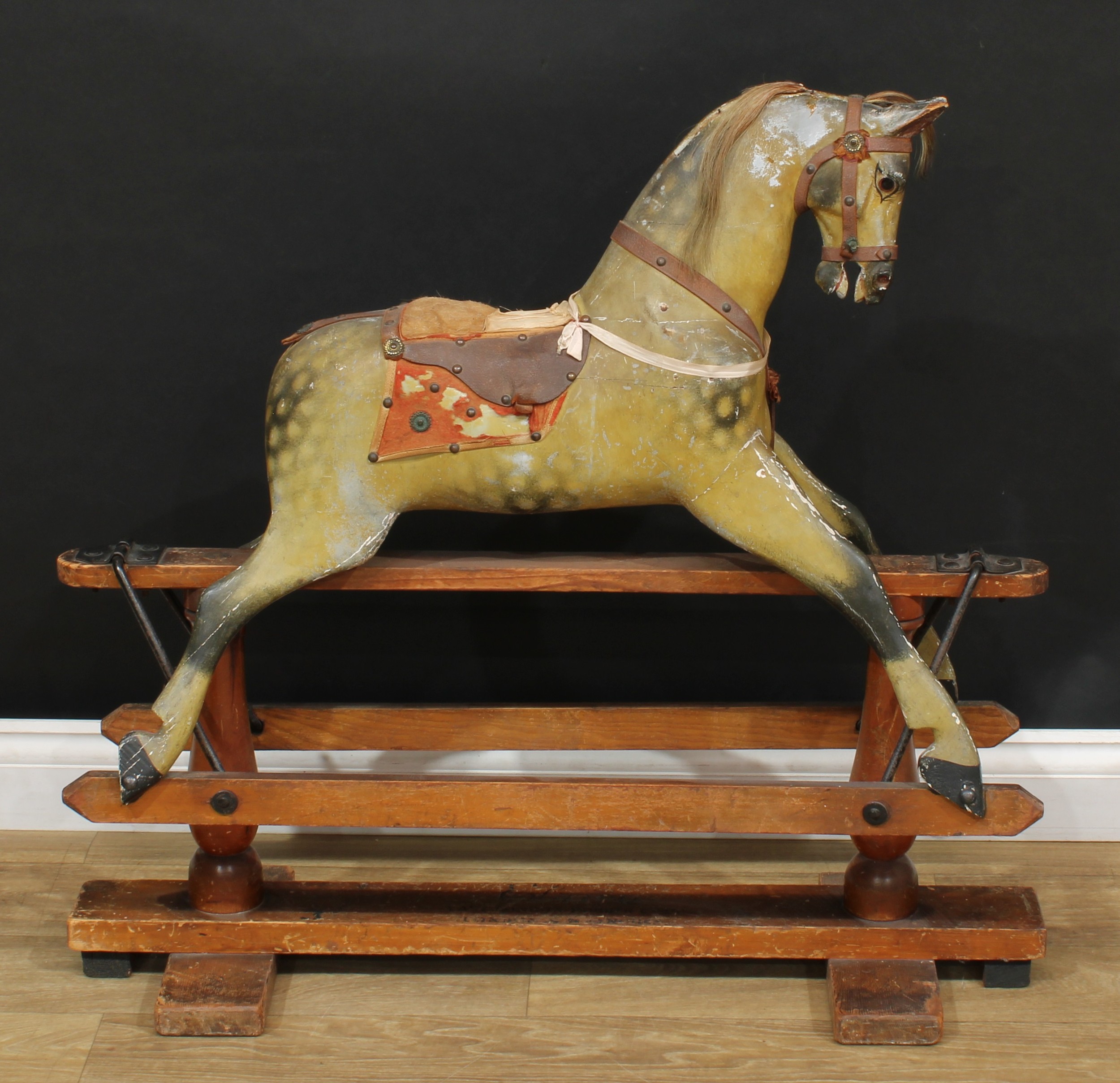 A late 19th/early 20th century English rocking horse of small proportions on safety stand, probably by F H Ayres (Frederick Henry Ayres, London), the carved, gesso and painted dappled horse with pricked ears, flared nostrils and open mouth revealing teeth, inset glass eyes and remnants of real horse hair mane, the body with outstretched legs on a safety stand, raised upon a pair of turned stand pillars and mounted with shaped rectangular type brackets with three bolts, trademark side rails with pointed ends, the stand base with various stencilled marks including 'EAST LONDON RUBBER CO LTD, LONDON & MANCHESTER, 3', 85cm high, 94cm long