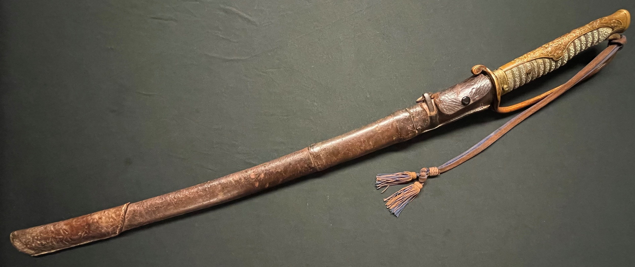 WW2 Imperial Japanese Army Officers Kyu Gunto Sword with single edged fullered blade 605mm in length. Tang is signed to both sides. Two holes to tang. Wire bound shark skin grip. Bronze decorative mounts. Company grade Officers knot. Scabbard has active service leather cover in place. Plus a green cotton cover. New to the market, war souvenir of a British Serviceman.