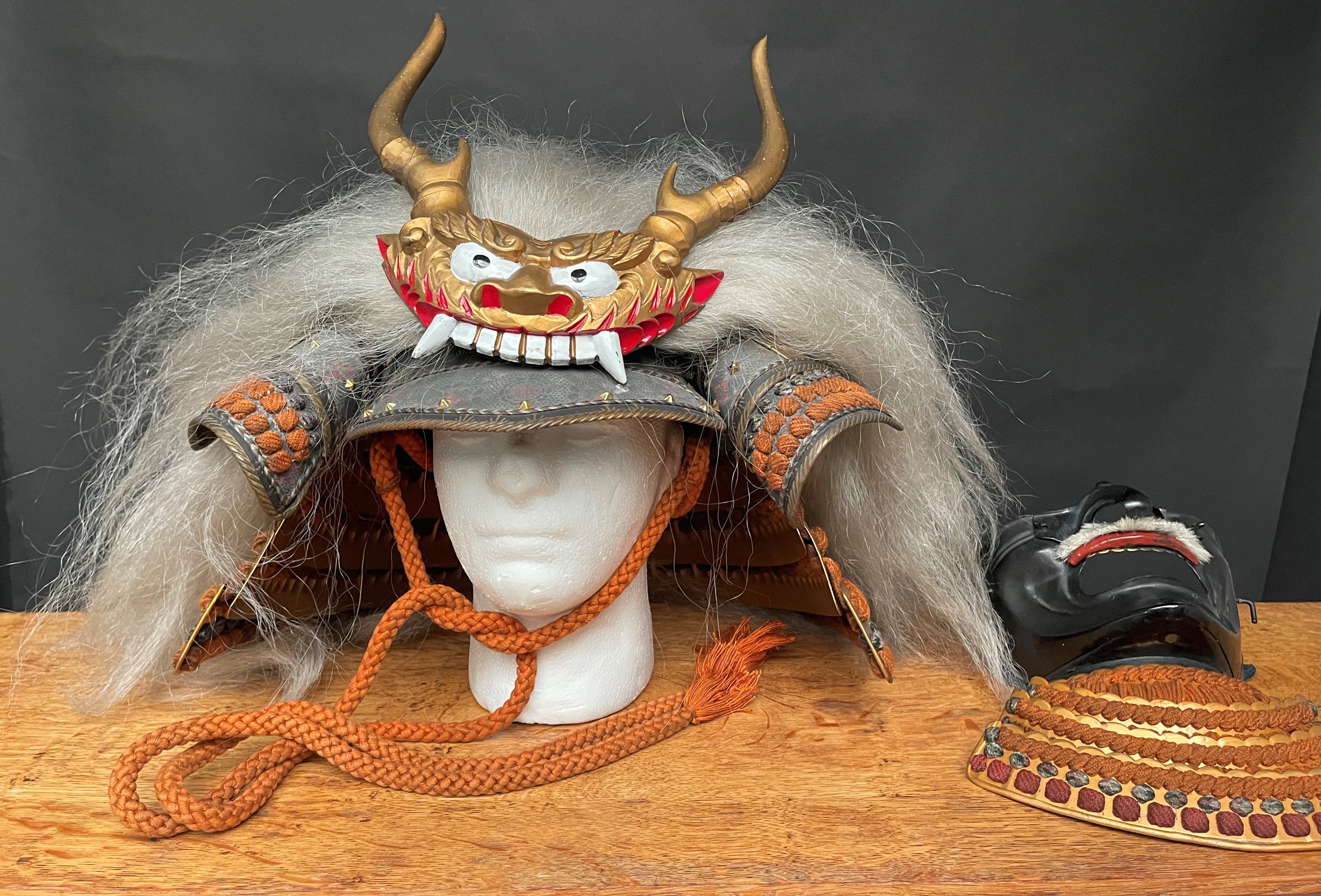 Japanese Samurai Warriors Kabuto Helmet, Faceplate, Greaves shin armour, brown fur shoes plus a black wooden slot together Sword stand, all contained in a black lacquered box with decorative brass mounts. Size width 65cm, height 57cm, depth 47cm.