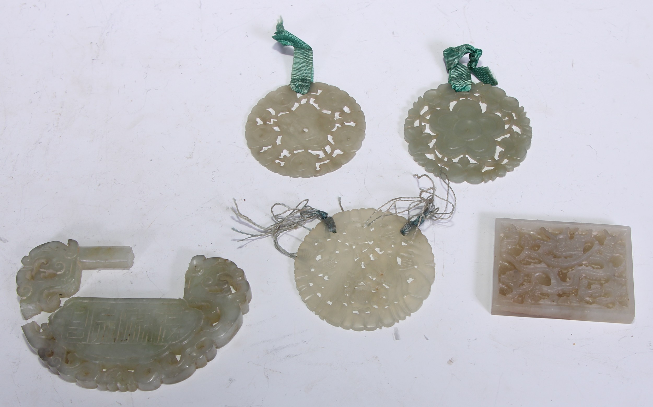 Three Chinese pale celadon/white nephrite disks, each carved with styalised flower heads, one with birds, remnants of silk tassels and ribbons, 5.5-6cm dia; a Chinese pale greenish-white nephrite plaque in the form of a lock plate (faults); a rectangular Chinese pale greenish-white nephrite plaque carved with a dragon, 5.75cm high (5).