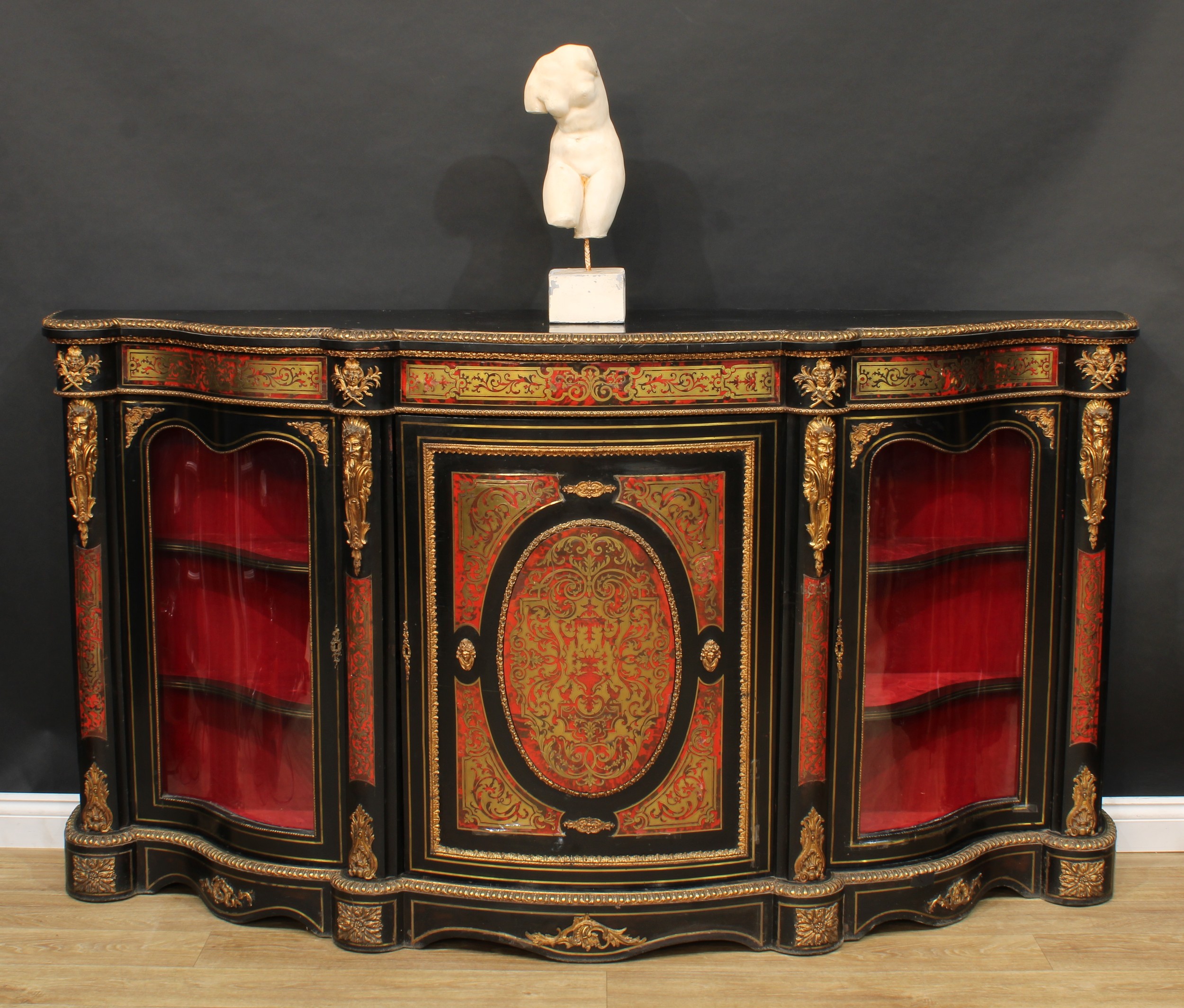 A 19th century gilt metal mounted Boulle and ebonised credenza, slightly oversailing serpentine top applied with egg-and-bell husk edge above a central panel door enclosing a shelf, flanked by glazed vitrine doors, each enclosing two further shelves, divided and flanked by herm brackets, shaped plinth base, 111cm high, 207cm wide, 49cm deep, c.1870