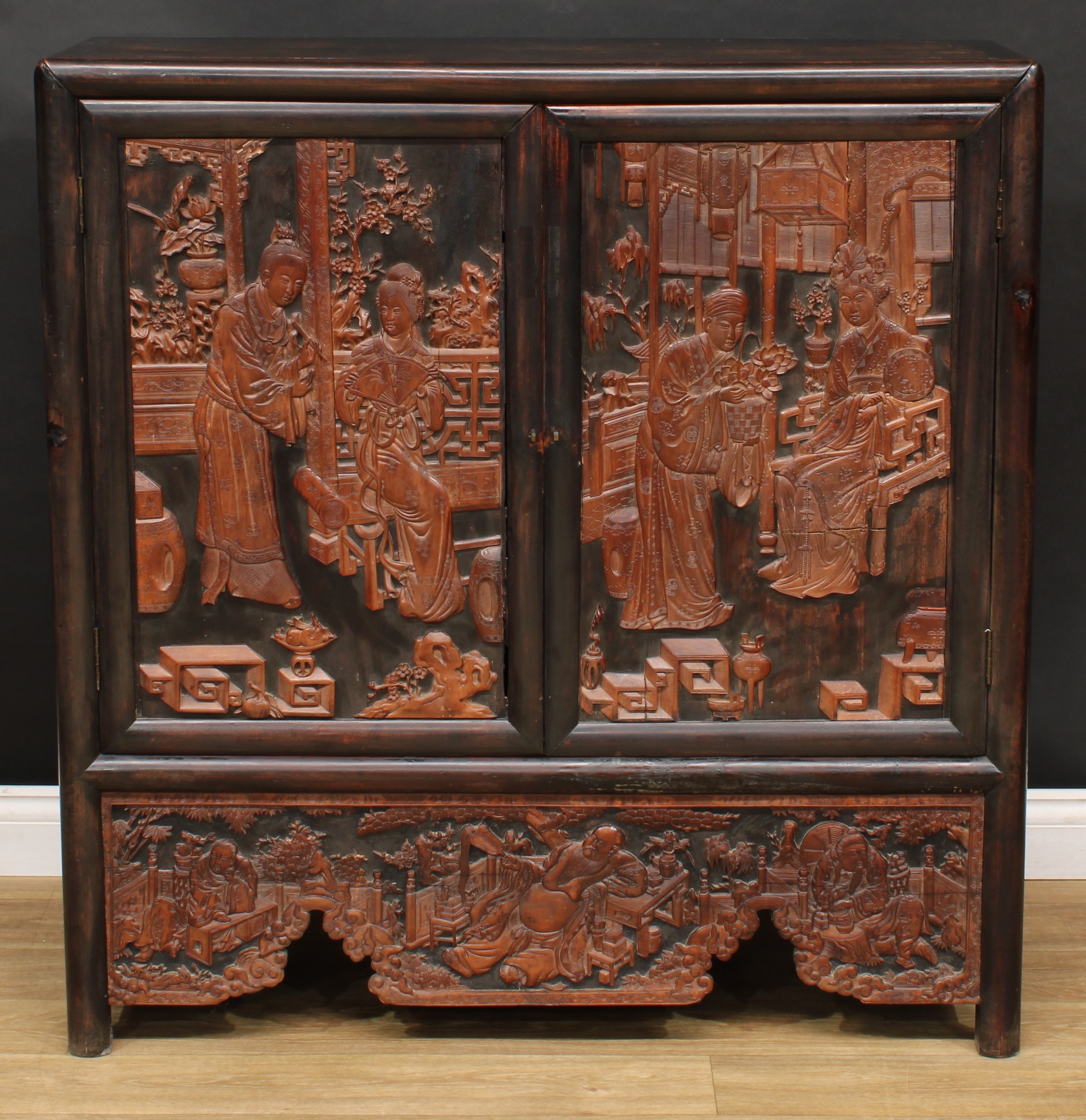 A Chinese hardwood side cabinet, flush rectangular top above a pair of panel doors carved and applied with ladies of the court and attendants within a pagoda amongst auspicious objects, enclosing two shelves, shaped apron carved and applied with Budai and other figures, 112cm high, 107cm wide, 37.5cm deep, early 20th century