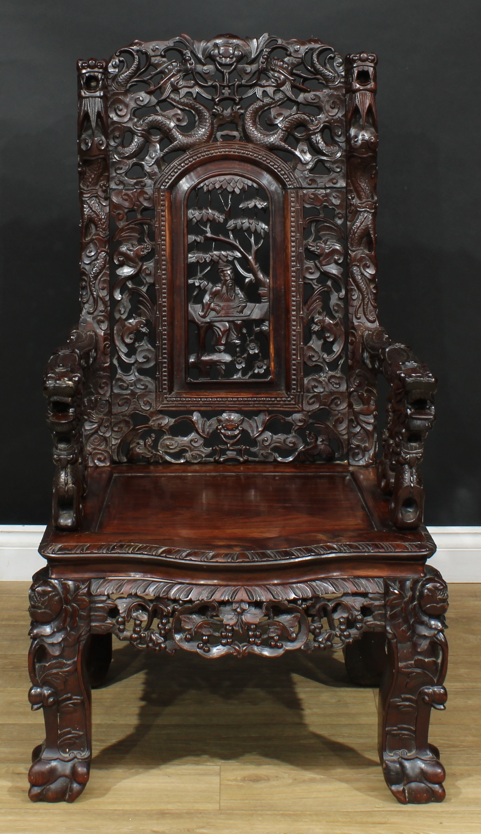 A substantial Chinese hardwood armchair, the back pierced and carved throughout with dragons, bats and scrolling clouds centred by an arched rectangular panel pierced and carved with a scholar beneath a tree, serpentine arms carved with a dragon, panel seat, shaped apron carved with fruiting vine, cabriole legs, ball and claw feet, 117.5cm high, 66.5cm wide, the seat 46cm wide and 46cm deep