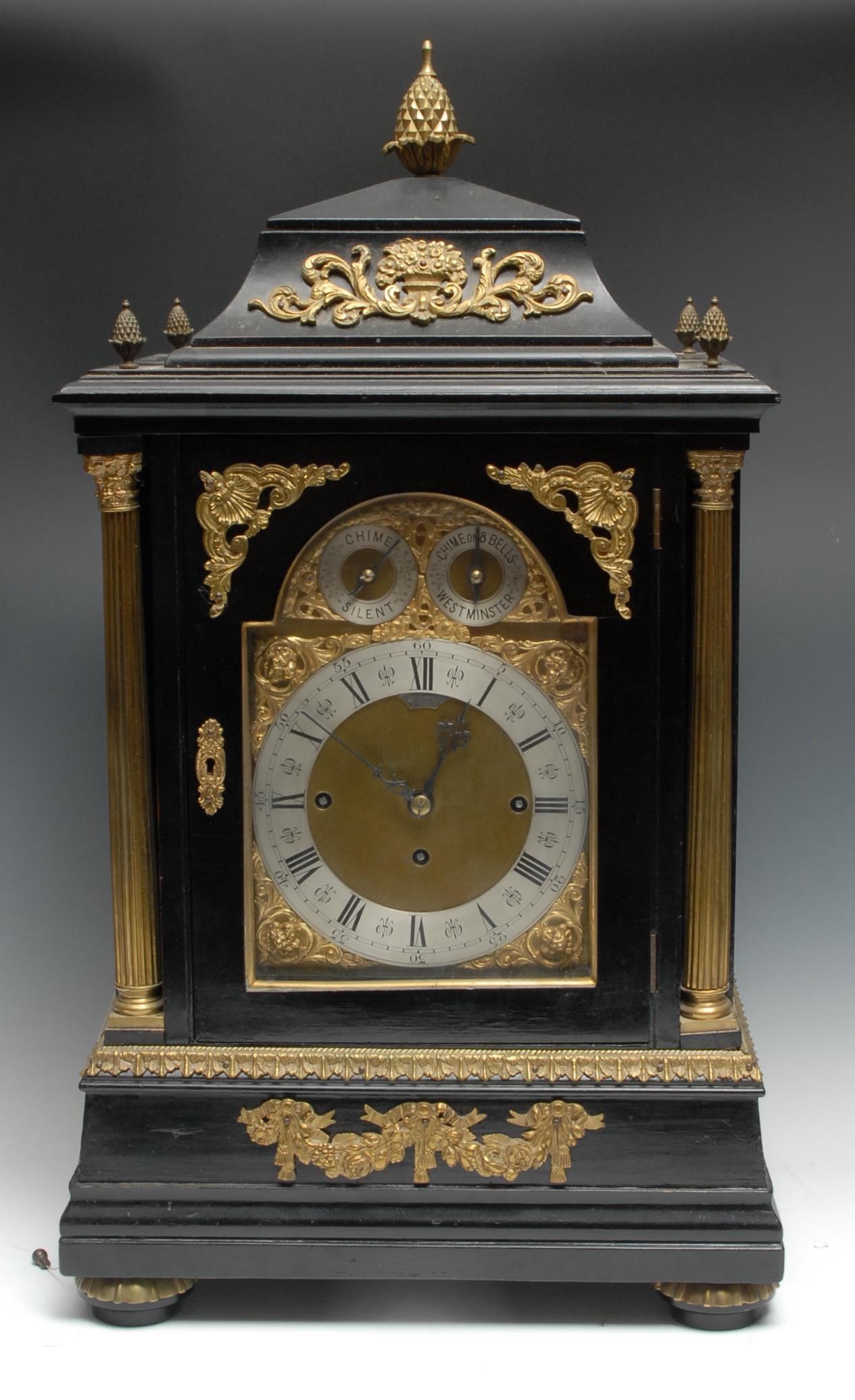 A substantial Victorian gilt-metal mounted ebonised repeating musical bracket clock, in the George II taste, 20cm arched brass dial with silvered chapter ring, inscribed with Roman and subsidiary Arabic numerals, Chime/Silent and Chime on 8 Bells/Westminster setting dials to arch, three-train movement striking on a gong, the caddy-top case with pine cone finials, flanked by a pair of Corinthian columns, fluted bun feet, 71cm high, c. 1880