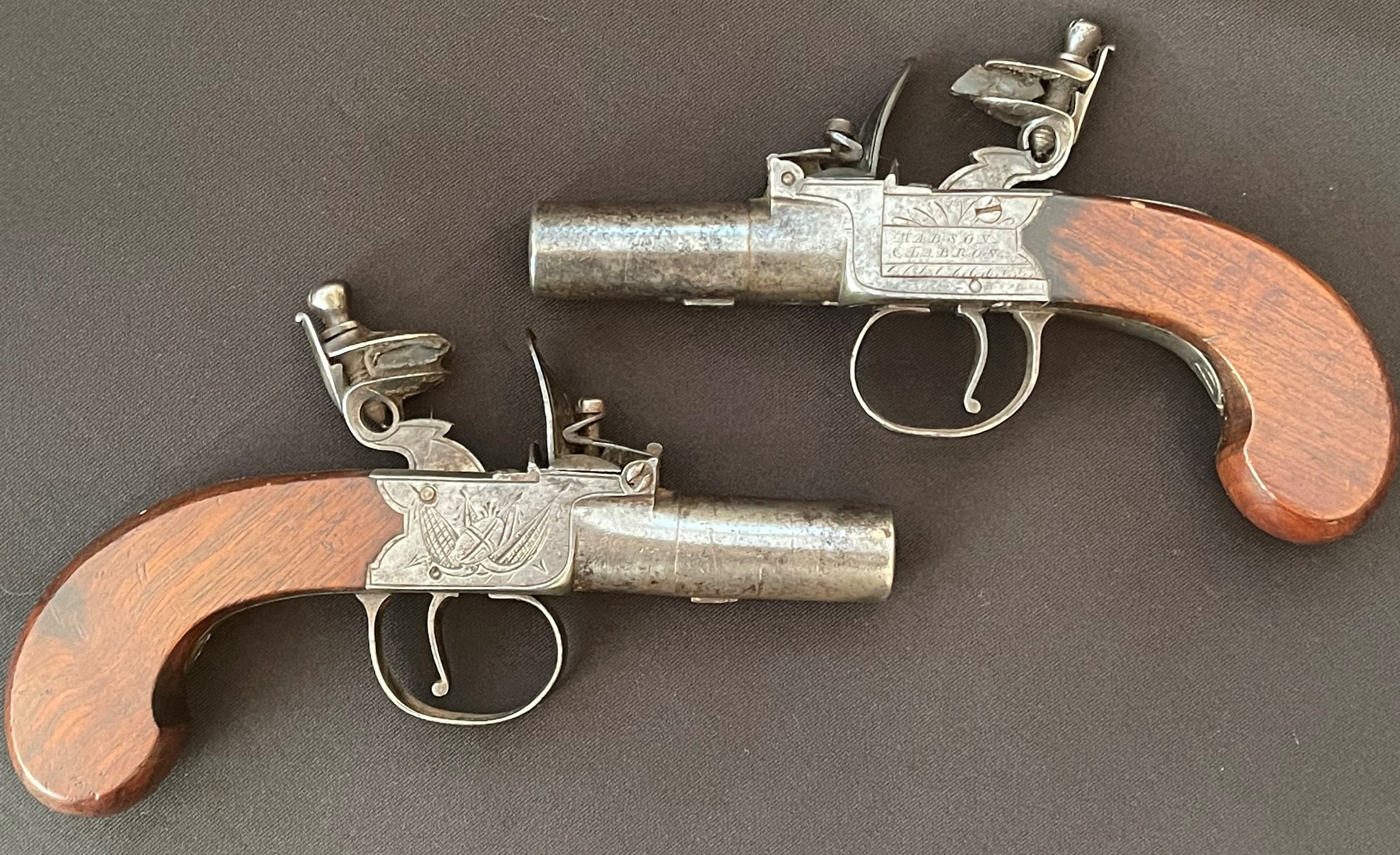 A matched pair of Flintlock Pocket Pistols by "Mabson & Labron". Both with with 55mm long screw on barrel. Bore approx. 12mm. Working A matched pair of Flintlock Pocket Pistols by "Mabson & Labron". Both wiaction which holds at both half and fully cocked positions. English proof marked. Maker marked to lock plate "Mabson & Labron" along with engraved piled arms motif. The maker was located in Birmingham and was in business under this trade name from 1822 until 1834.  Walnut grips. Overall length 150mm. Section 58 (2) no licence required. (2)
