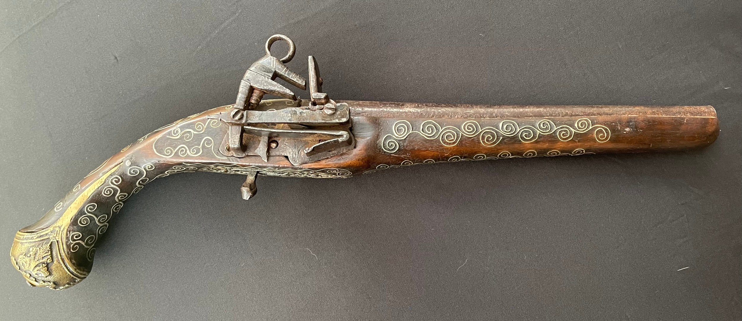 Balkan style Flintlock Pistol with 270mm long barrel. Approx. 13mm Bore. Working action. Decorated stock inlaid with scrolls of silver overall. Cast decorative brass butt plate. Jaws also inlaid with silver wire. Overall length 460mm. No makers marks or proof marks. Section 58 (2) no licence required.
