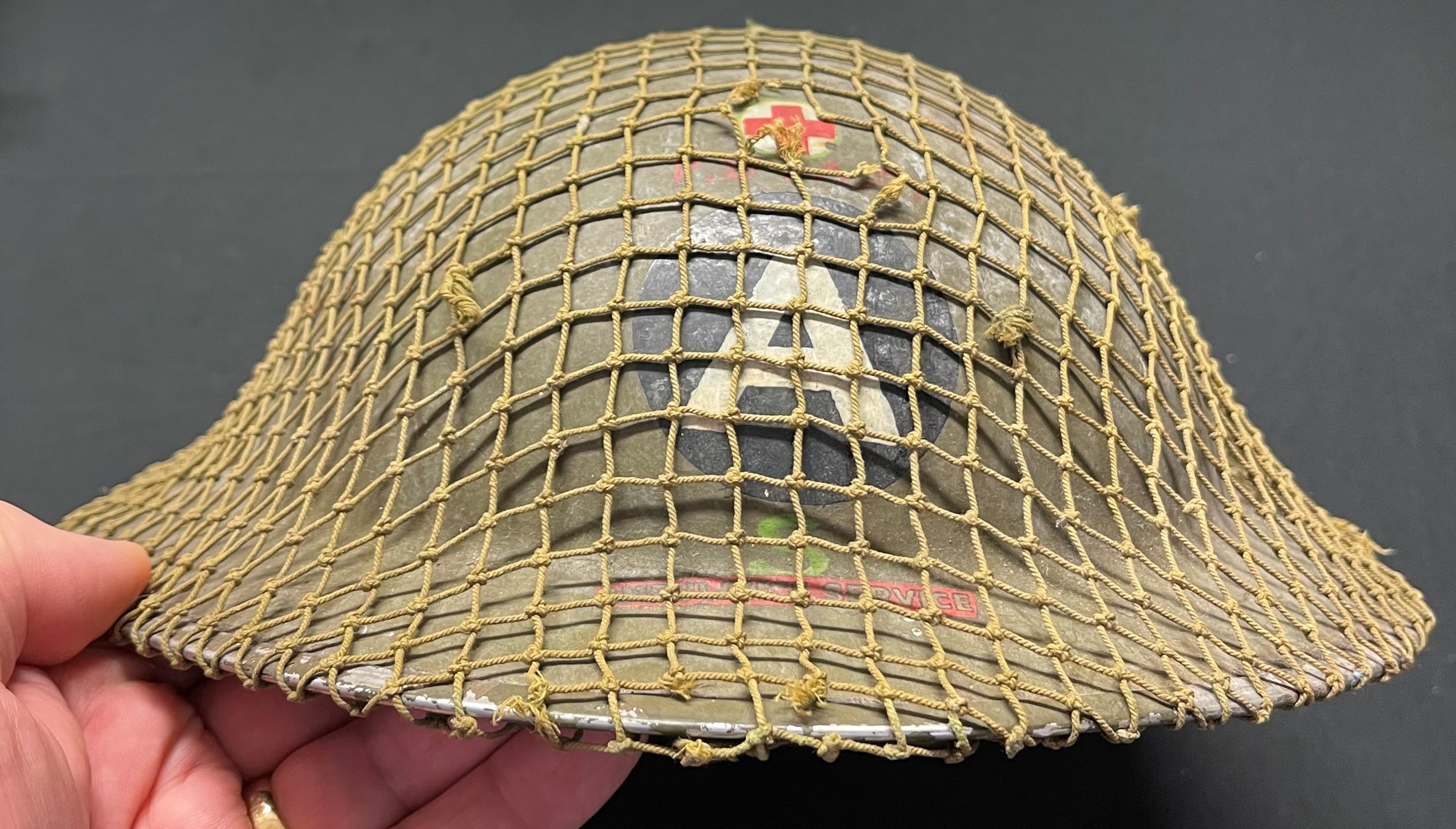 WW2 Rare British Home Front Steel Helmet for the Peak District Ambulance Service. Insignia to the front of the helmet is a mixture of hand painted Red Cross, "A" for Ambulance, red PDAS, green letter "S" along with a decal for the wording "Ambulance Service". The helmet is covered with it's original camo netting cover. To the inside is a panel painted in grey with the owners initials "FS" painted in black. The chinstrap has two period repairs done in canvas cloth. This helmet is new to the market never having been in a collection before. It is being sold by the family of the original owner. Also included is an original photo of the owner, Mr Fred Stansfield and two showing the unit's ambulances.