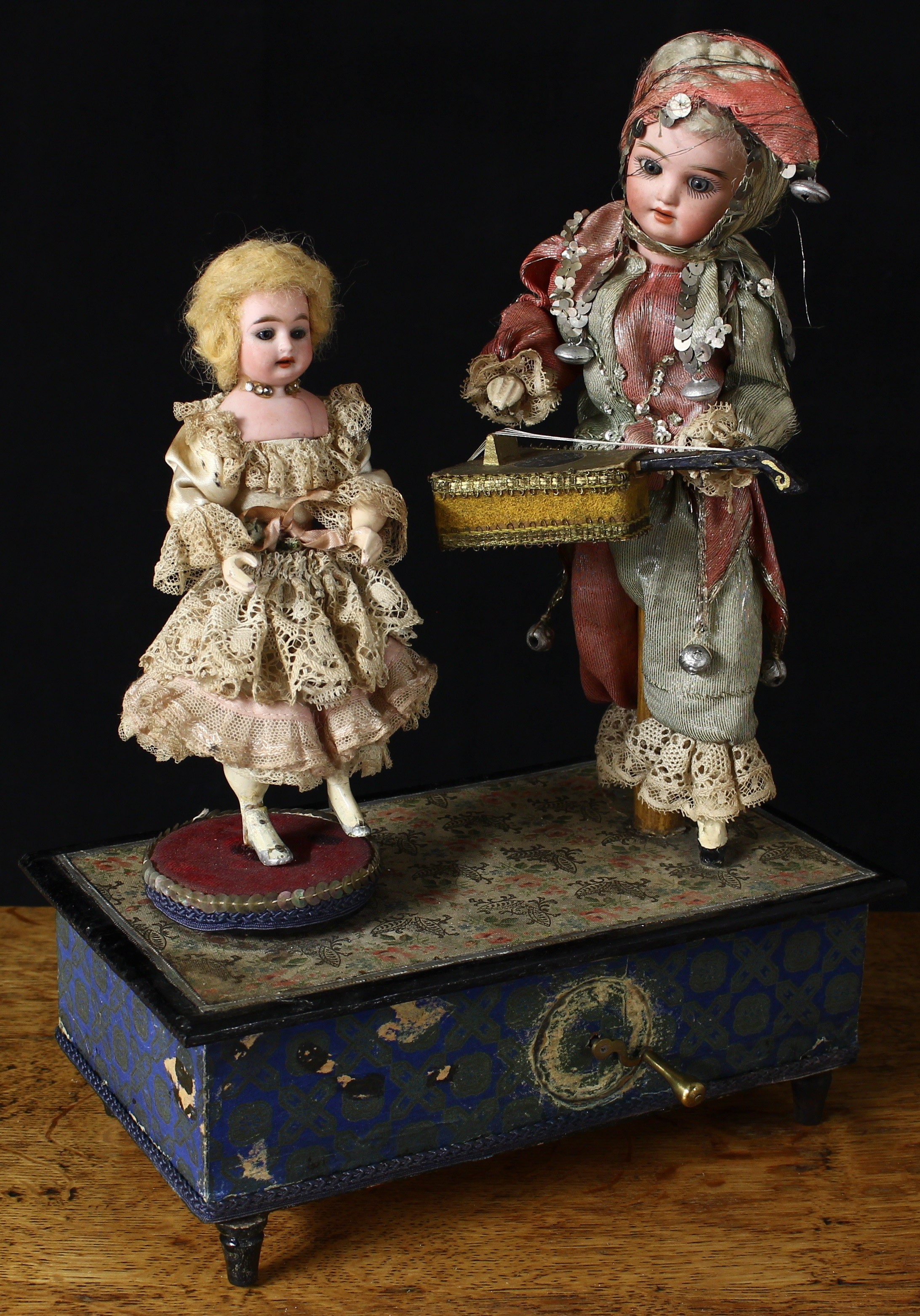 A late 19th century/early 20th century German novelty musical automaton group, ‘The Musician and Dancer', comprising a Schoenau & Hoffmeister/Porzellanfabrik Burggrub (Germany) bisque head doll, dressed in costume and holding a lute, inset fixed blue glass eyes, painted features including fine eyelashes and brown feathered eyebrows, open mouth revealing upper teeth, impressed marks, the bisque shoulder head dancer, dressed in a silk dress with lace detail, inset fixed blue glass eyes, painted features including fine eyelashes and brown feathered eyebrows, she stands upon a circular platform, the group mounted upon a rectangular shaped wooden box applied with printed paper, brass crank handle, the box fitted with a musical mechanism which when wound plays Boccherini’s string quintet Minuet, the musician moves the head from side to side and the free arm moves back and forth above the lute whilst the dancer rotates, 37cm high, 25cm wide, 15cm deep