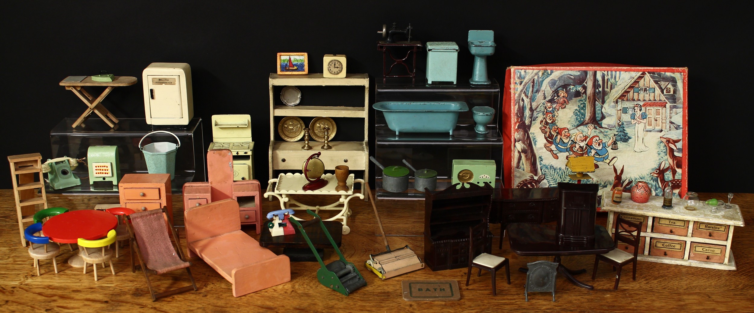 A collection of dolls house furniture, curios and accessories, first-half 20th century and later comprising a Kleeware part dining room suite including dining table, two chairs, sideboard, dresser and wireless radio; a wooden constructed shop display unit, comprising an arrangement of six drawers, each applied with pins for drawer handles and paper labels applied for Cinnamon, Tea, Raisins, Spicery, Coffee and Cacao; a Dol-Toi painted wooden refrigerator with a single bottle of milk; a Dol-Toi collapsible wooden ironing board; a DCMT Crescent lead cooker; other accessories, some lead and a set of Snow White and the Seven Dwarfs picture puzzle blocks, boxed etc (quantity)