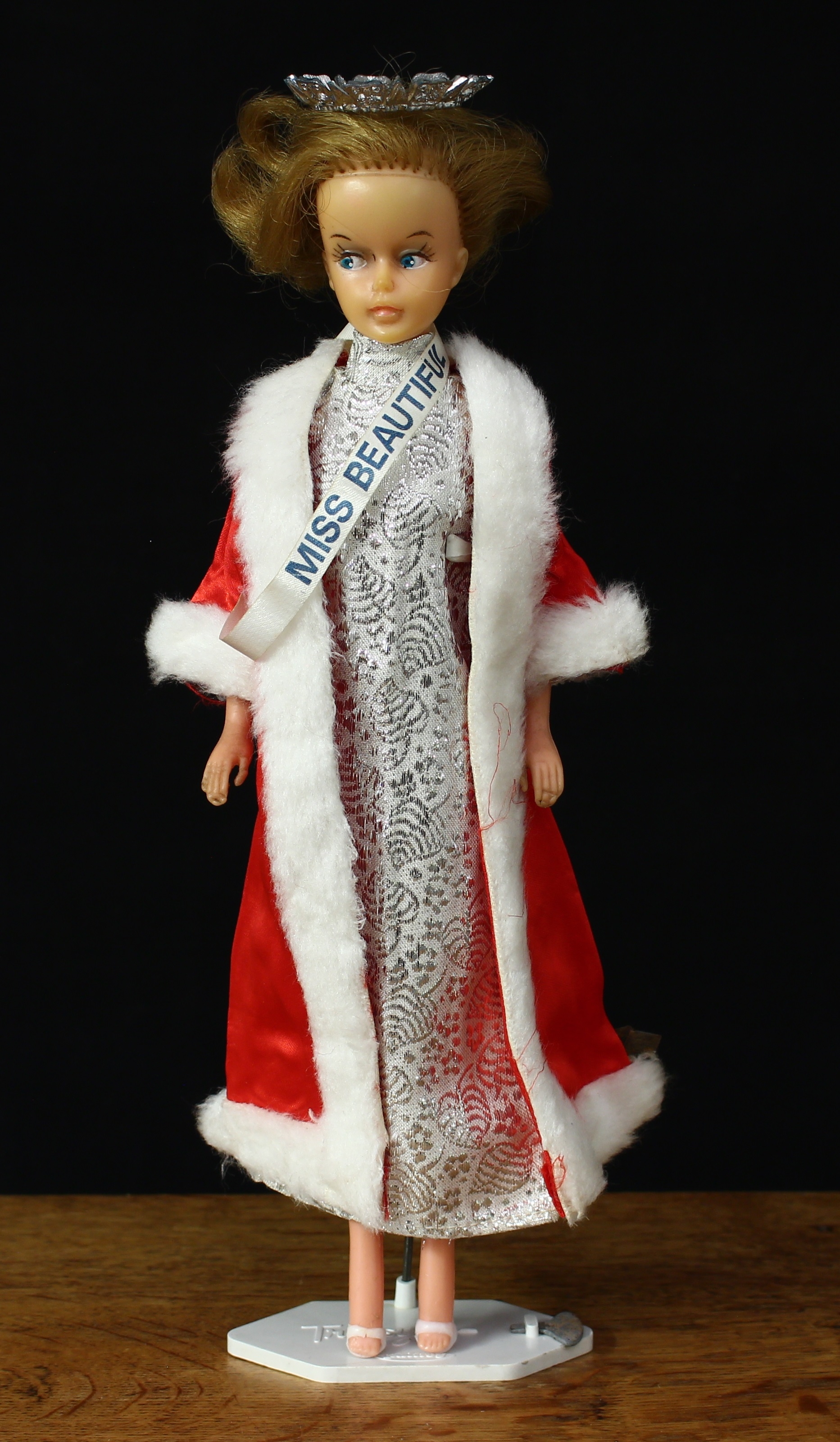 A 1960’s Palitoy Tressy doll, mid blonde hair, side glancing blue eyes, wearing a beauty pageant outfit with ‘MISS BEAUTIFUL’ sash and crown, white high heel shoes, 30cm high, unboxed with key and ‘Tressy’ display stand