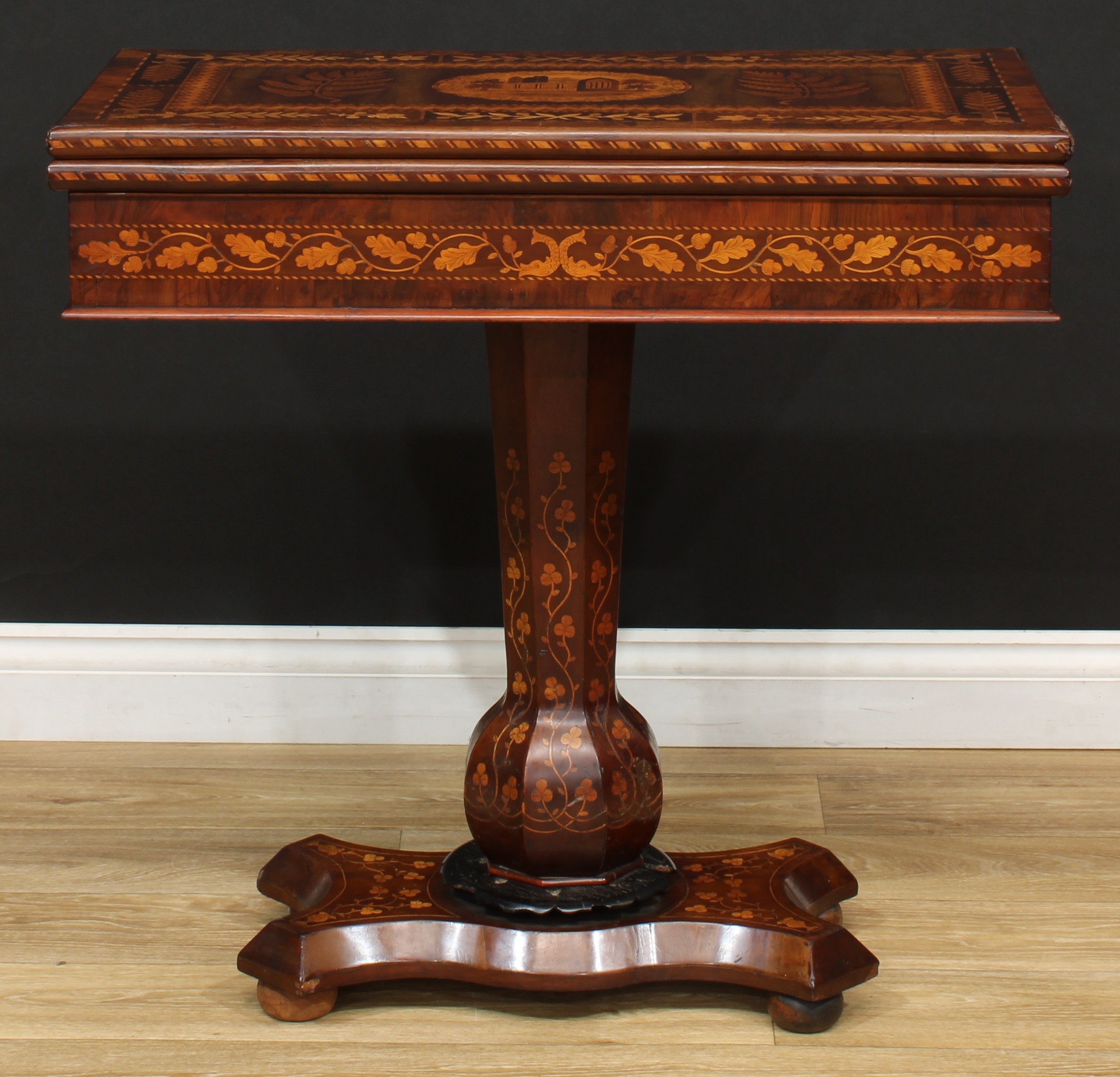 A 19th century Irish Killarney marquetry and yew games table, hinged rectangular top inlaid with an oval reserve of Muckross Abbey, County Kerry, Ireland, bordered by trailing sprays of seamróg shamrocks and outlined with barber-pole stringing, herringbone parquetry bands and stiff leaves, enclosing playing surfaces for chess, backgammon and cribbage and further architectural landscapes, possibly Ross castle and Glena cottage, above a deep frieze inlaid with oak leaves and acorns, panelled 'garlic-mouth' column, serpentine rectangular base, bun feet, 77cm high, 79.5cm wide, 42cm deep, each chess square 4cm, c.1850