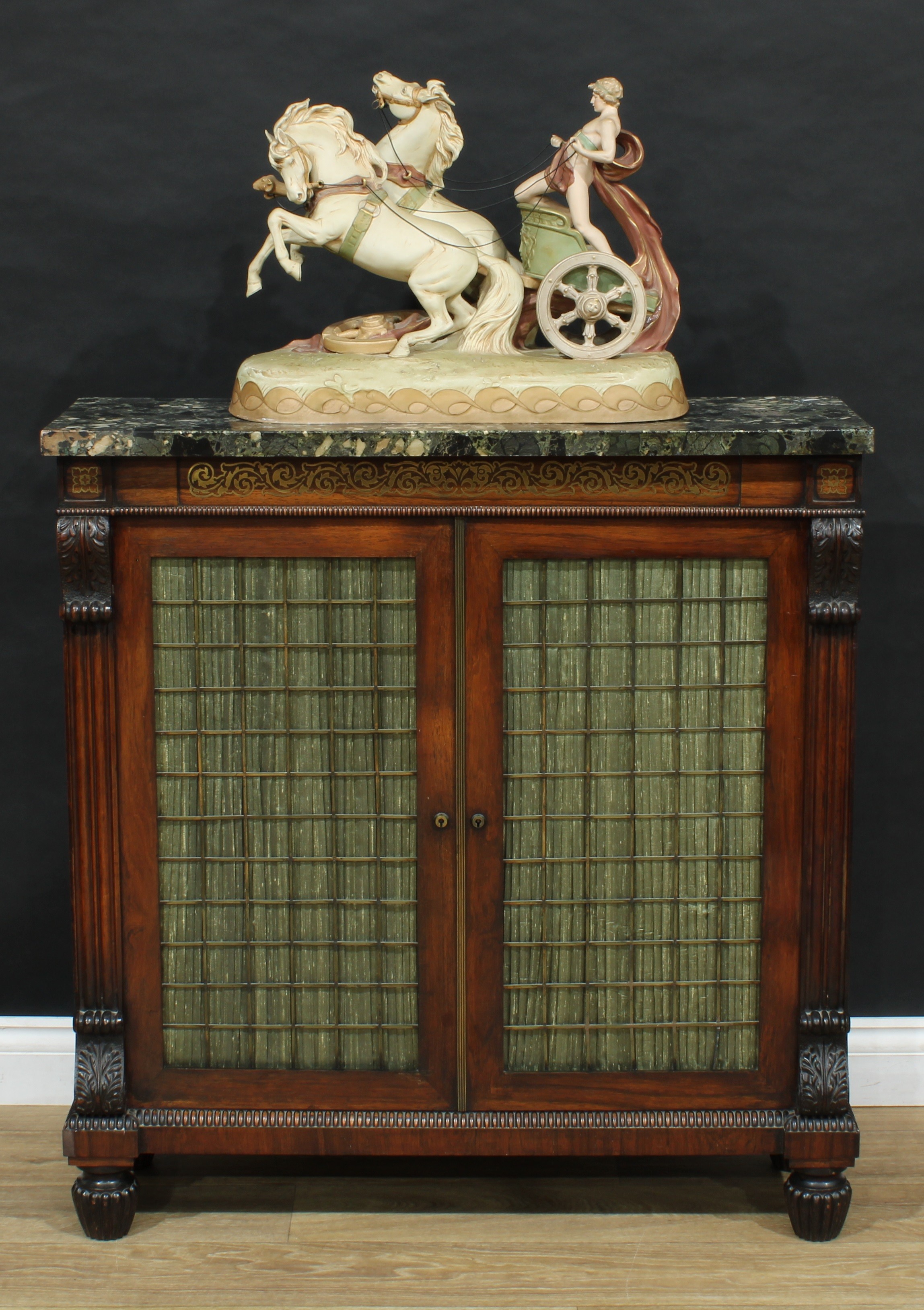 A Regency rosewood and brass marquetry library pier cabinet, attributed to Gillow of Lancaster and of small and neat proportions, rounded rectangular marble top above a deep frieze and a pair of rectangular doors, the brass grilles lined in pleated silk and enclosing adjustable shelves, flanked by acanthus scroll brackets, fluted bun feet, 96.5cm high, 92cm wide, 30.5cm deep, c.1820