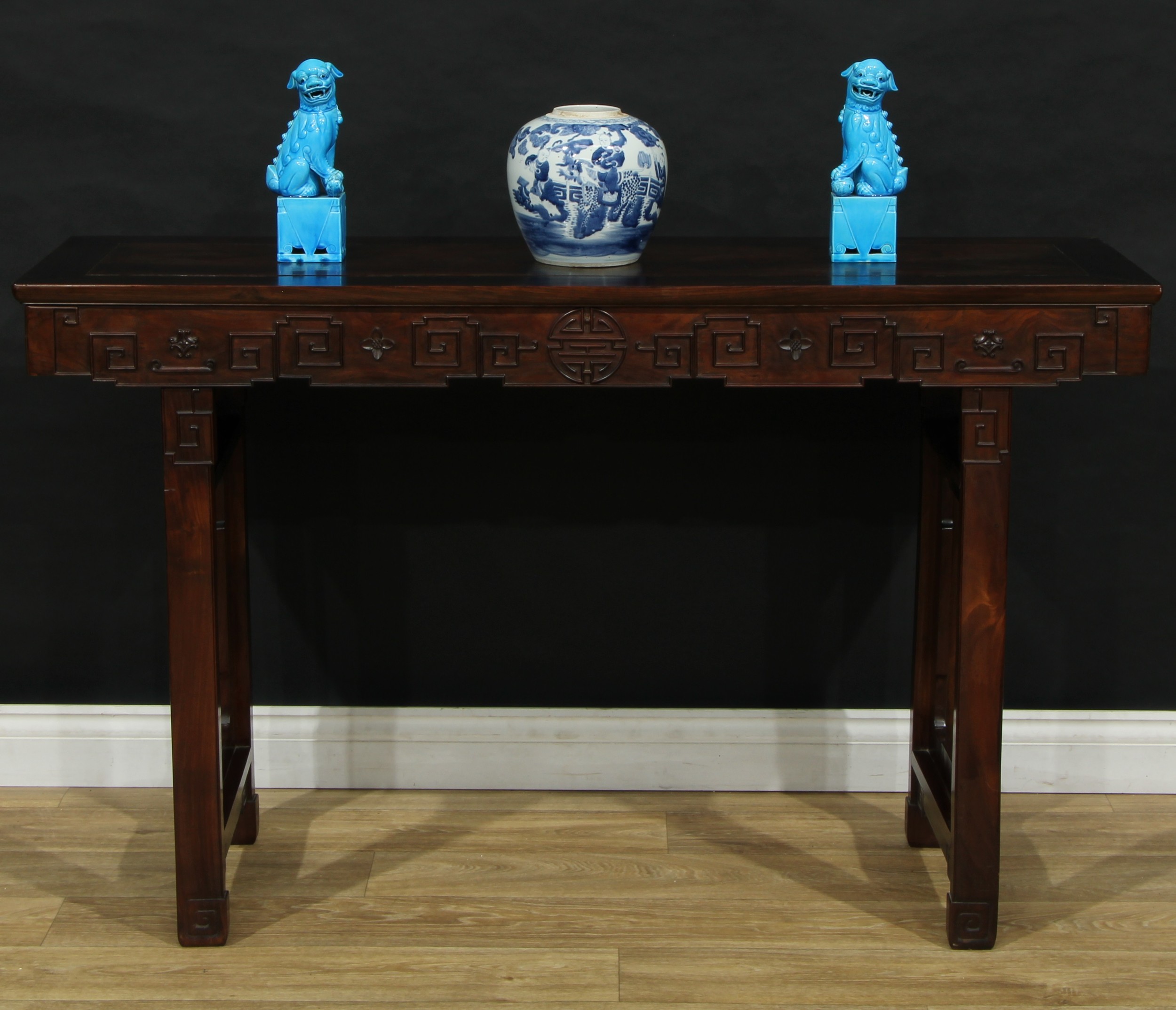 A Chinese hardwood altar table, rectangular panel top above a shaped frieze carved with double-happiness, angular scrolls and flowerheads, trestle underframe, 84cm high, 139cm wide, 42.5cm deep, c.1900