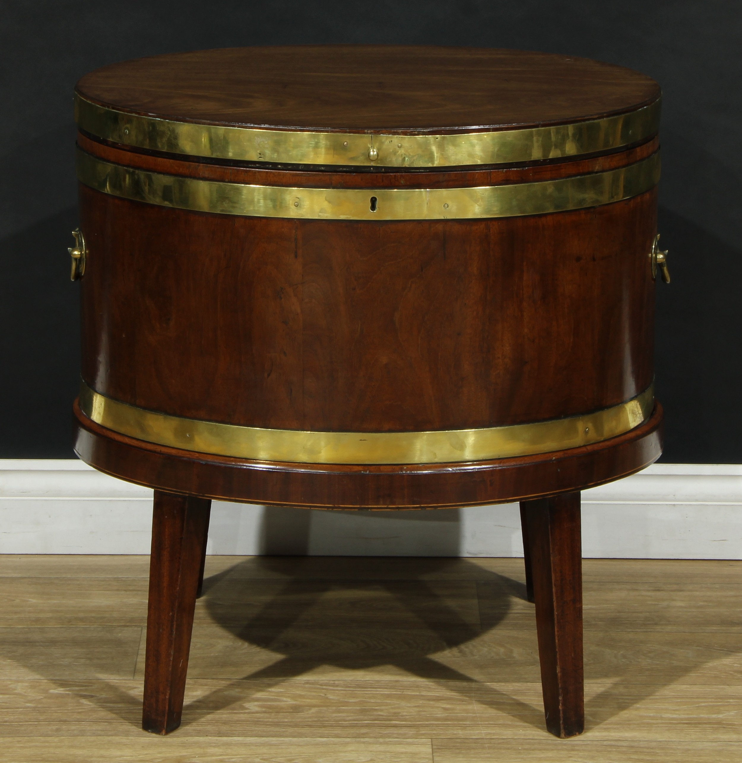 A George III brass-bound mahogany oval cellarette, hinged cover, zinc liner, swan neck carry handles to sides, tapered square legs, 67.5cm high, 66cm wide, 51cm deep, c.1780