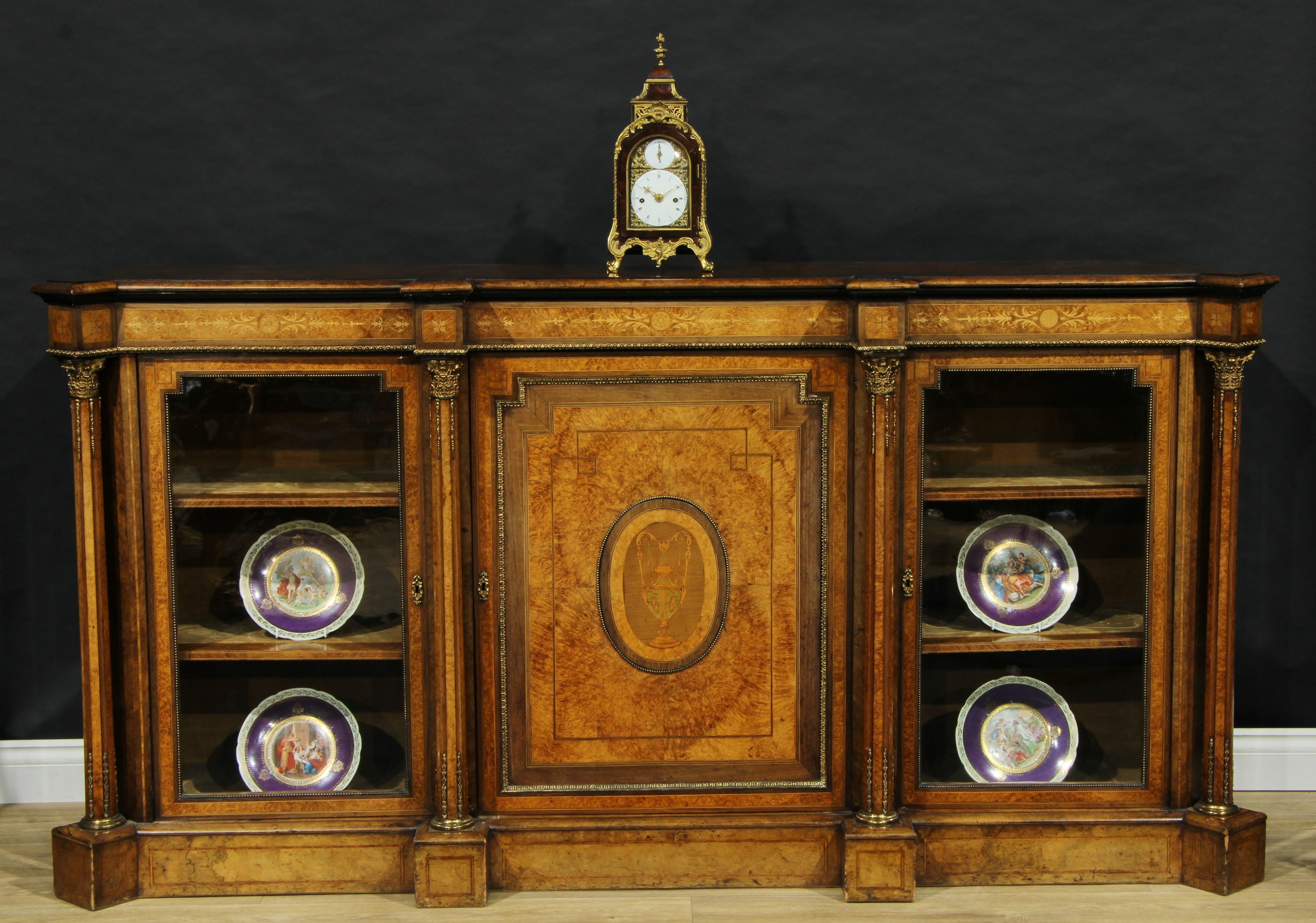 A Victorian gilt metal mounted burr walnut and marquetry credenza, moulded top above a rectangular panel door centred by an oval reserve inlaid with a Classical urn, flanked by a pair of glazed vitrine doors, crossbanded and outlined throughout with boxwood and ebony stringing, the projecting angles with Corinthian columns, plinth base, 105cm high, 202cm wide, 48cm deep, c.1870