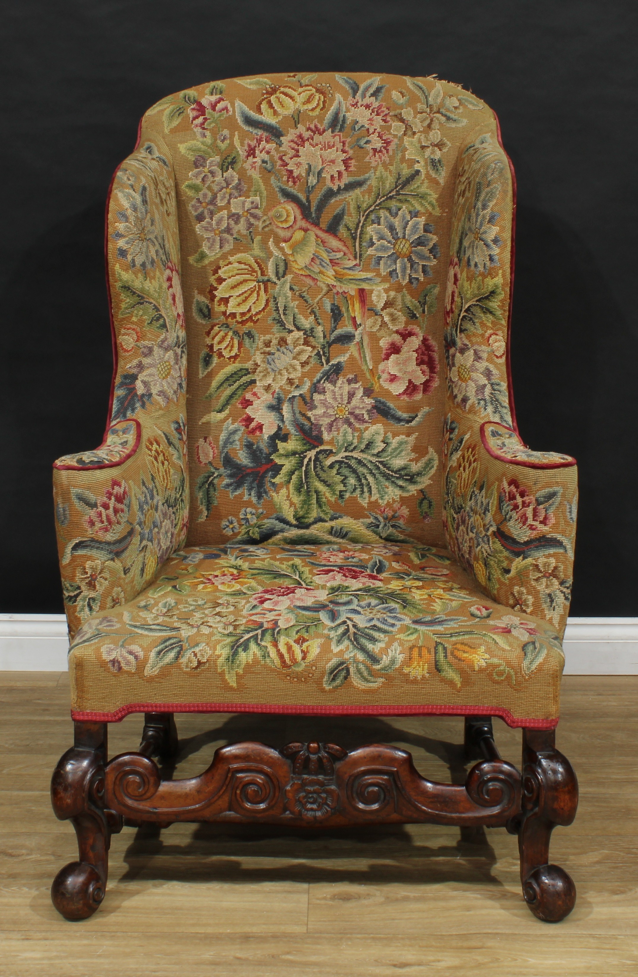 A 19th century wingback armchair, in the William and Mary taste, shaped back, outswept arms, stuffed-over tapestry upholstery, brightly worked with a parrot and flowers, shaped front rail and forelegs carved with scrolls, 122cm high, 79cm wide, the seat 47cm wide and 52cm deep