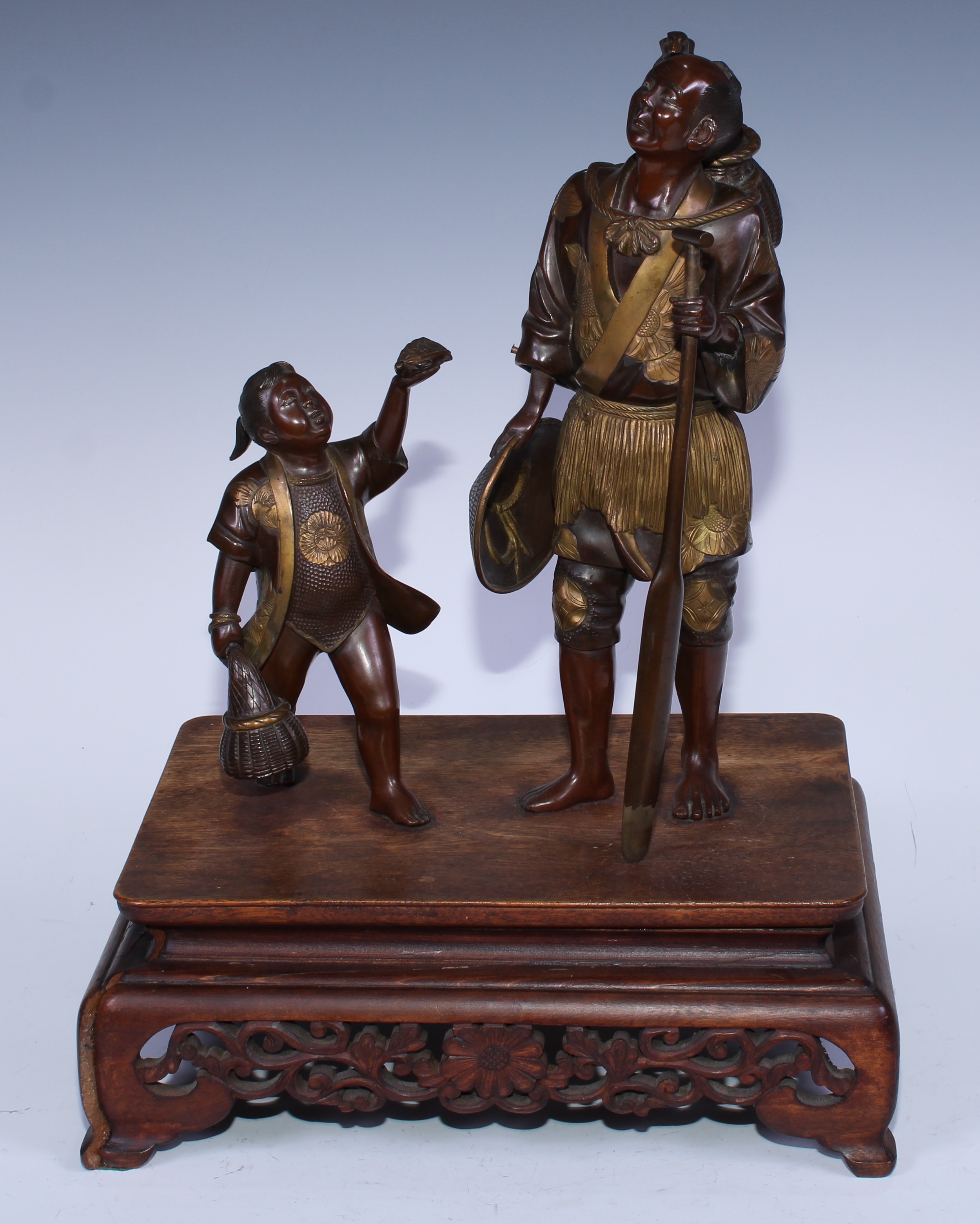 A Japanese brown and gilt patinated bronze group of a Cormorant fisherman and boy, he stands holding an oar with the bird resting on the creel on his back, the boy offers a terrapin, hardwood base, 40cm high overall