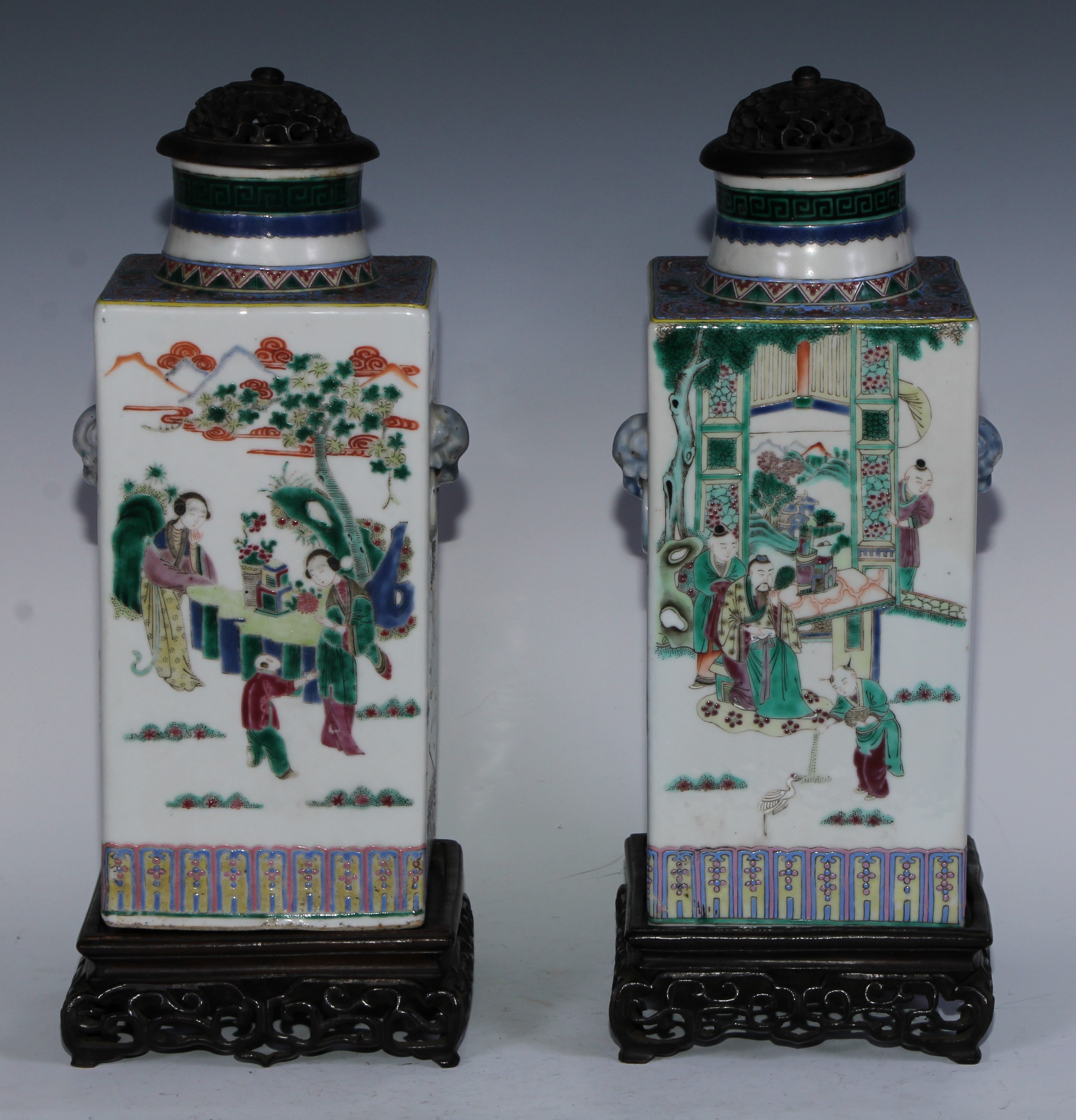 A pair of Chinese square vases, painted in polychrome in the famille verte and rose palette with figures of the court and monumental landscapes, moulded temple lion mask ring handles, 30.5cm high, four character marks, 19th century, carved hardwood covers and stands
