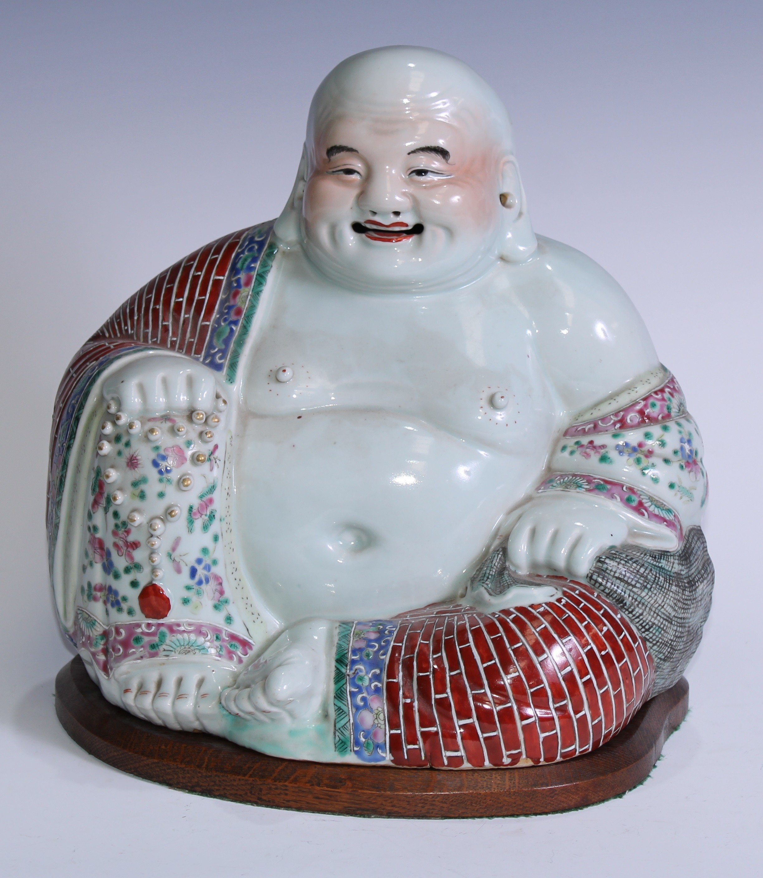 A Chinese porcelain model, of Budai, seated, smiling, brightly painted in polychrome enamels, 29cm high, impressed mark, early 20th century, wooden stand