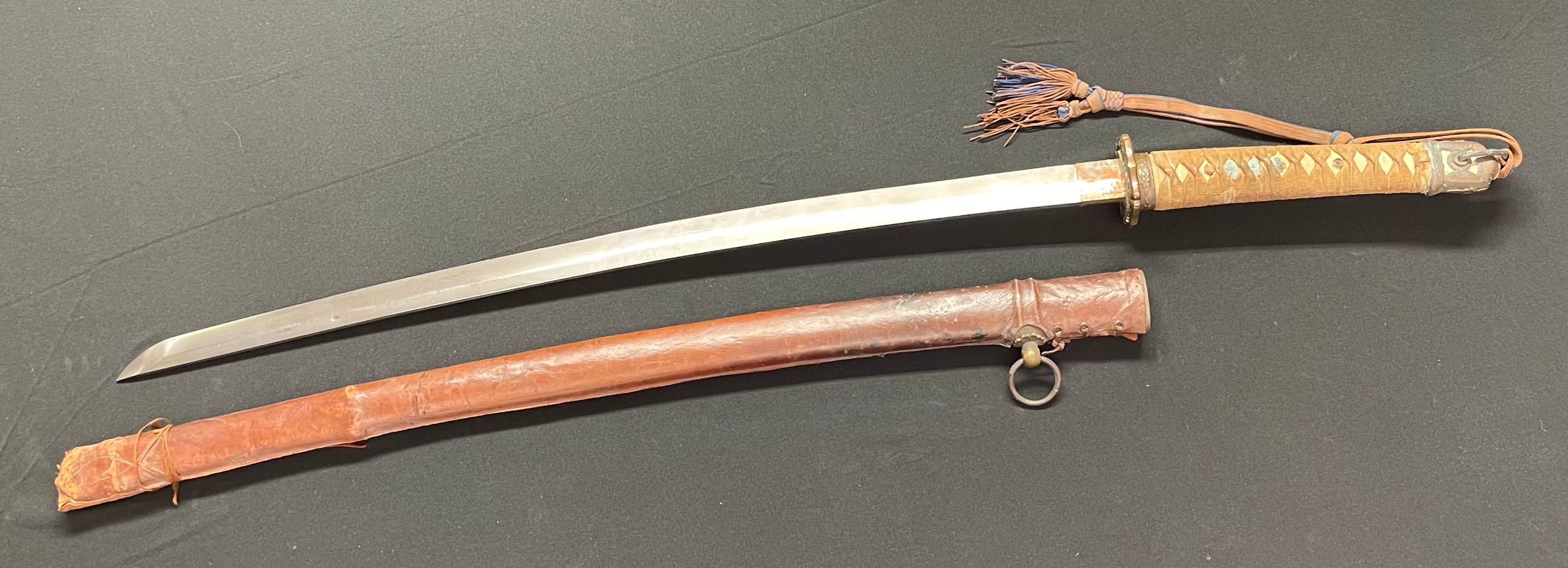 WW2 Imperial Japanese Army Officers Shin-Gunto Katana Sword with 695mm long blade. Grip has original binding in place with Menuki beneath. Overall length 950mm. Machine made blade with no visible Hamon line. "49" stamped on the Habaki. Working release catch. Complete with original company grade officers blue/brown knot and active service leather cover for the Saya scabbard.