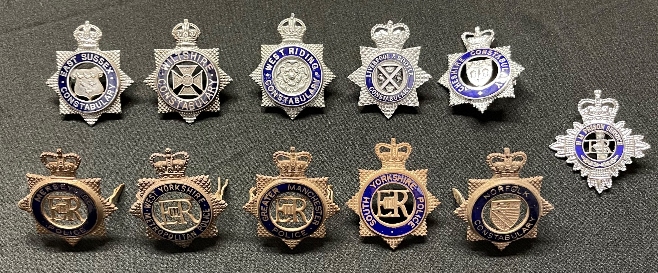 A collection of Police Officers cap badges to include: Kings Crown West Riding Constabulary: Kings Crown Wiltshire Constabulary: Kings Crown East Sussex Constabulary: Queens Crown Norfolk Constabulary: Queens Crown Merseyside Police: Queens Crown South Yorkshire Police: Queens Crown Greater Manchester Police: Queens Crown West Yorkshire Metropolitan Police: Queens Crown Liverpool & Bootle Constabulary: Queens Crown Cheshire Constabulary: Queens Crown HM Prison Swervice. (11)
