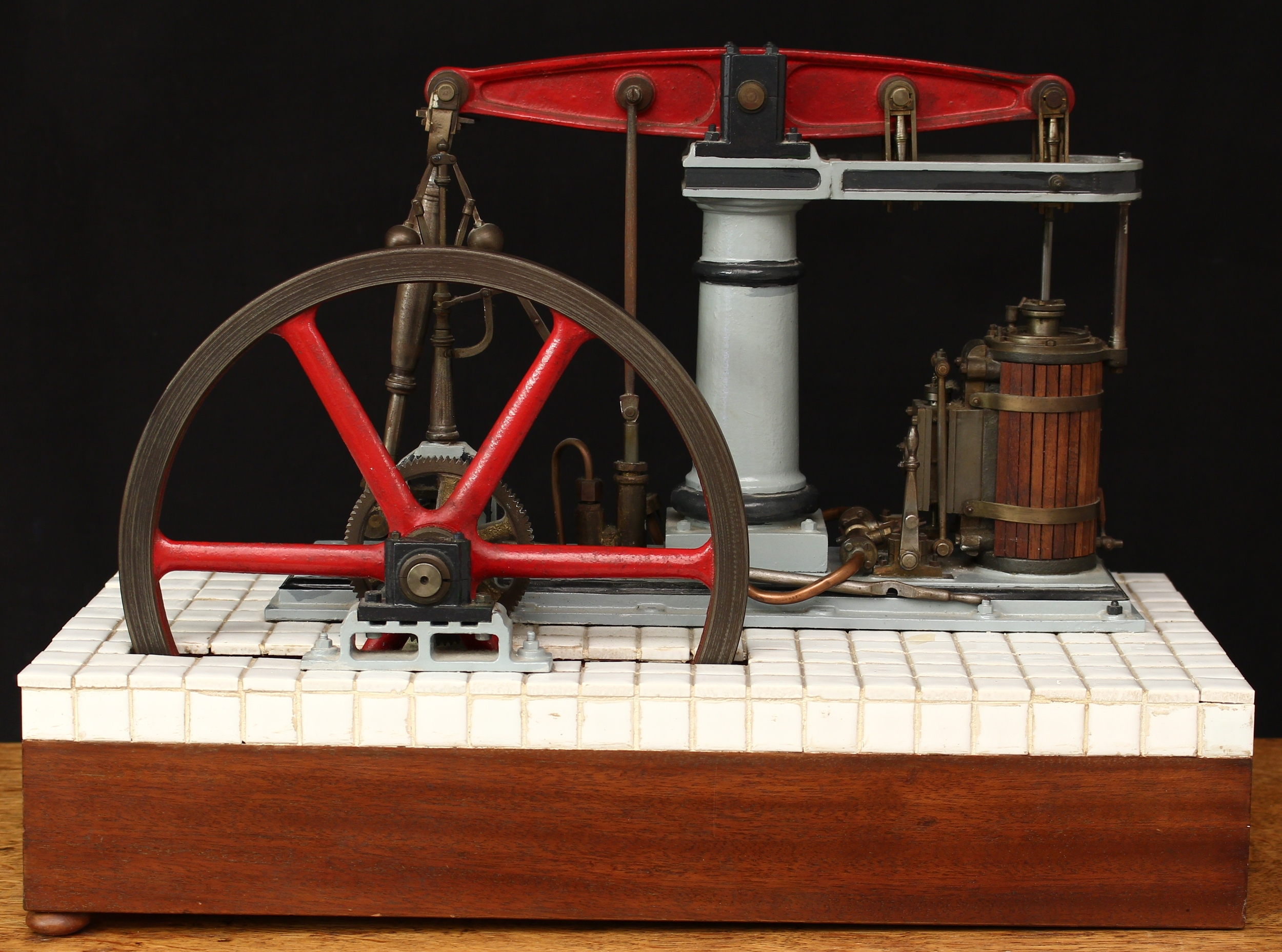 A live steam horizontal beam engine, the painted and turned central column supporting the beam, single wooden lagged cylinder, 9" diameter six spoke flywheel painted in red, mounted on a rectangular wooden plinth partially covered in small white ceramic tiles, 34cm high, 44.5cm wide, 22cm deep