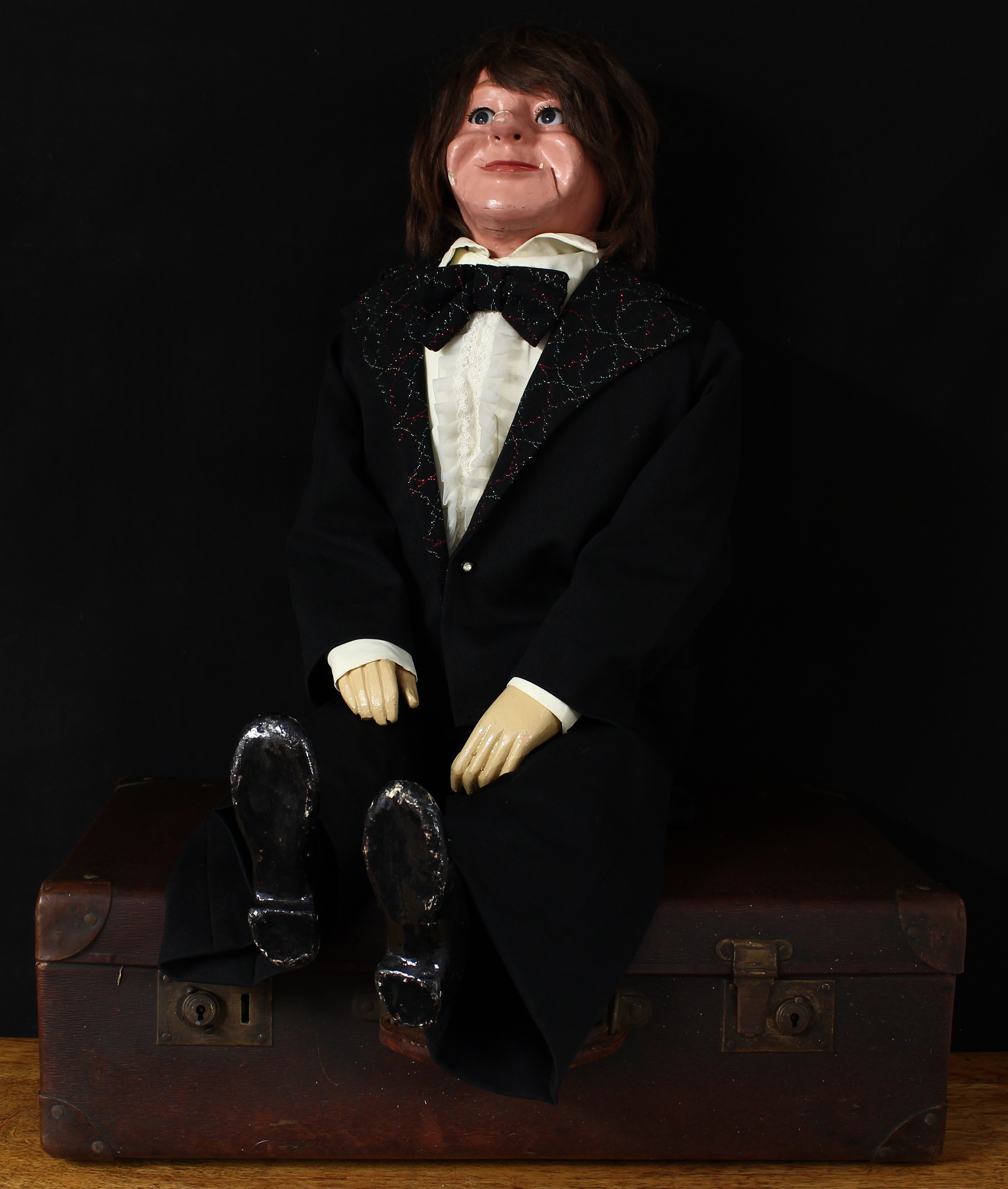 Ventriloquism/Ventriloquy – a mid-20th century ventriloquists dummy in the form of a gentleman, wearing a black dinner suit, the painted papier mâché head with mechanisms for the inset side glancing blue glass eyes and mouth piece, long brown wig, carved and painted wooden hands, the lapels on the black dinner jacket picked out with coloured threads and matching bow tie, white ruffled shirt and black trousers, 105cm high (suitcase not included)