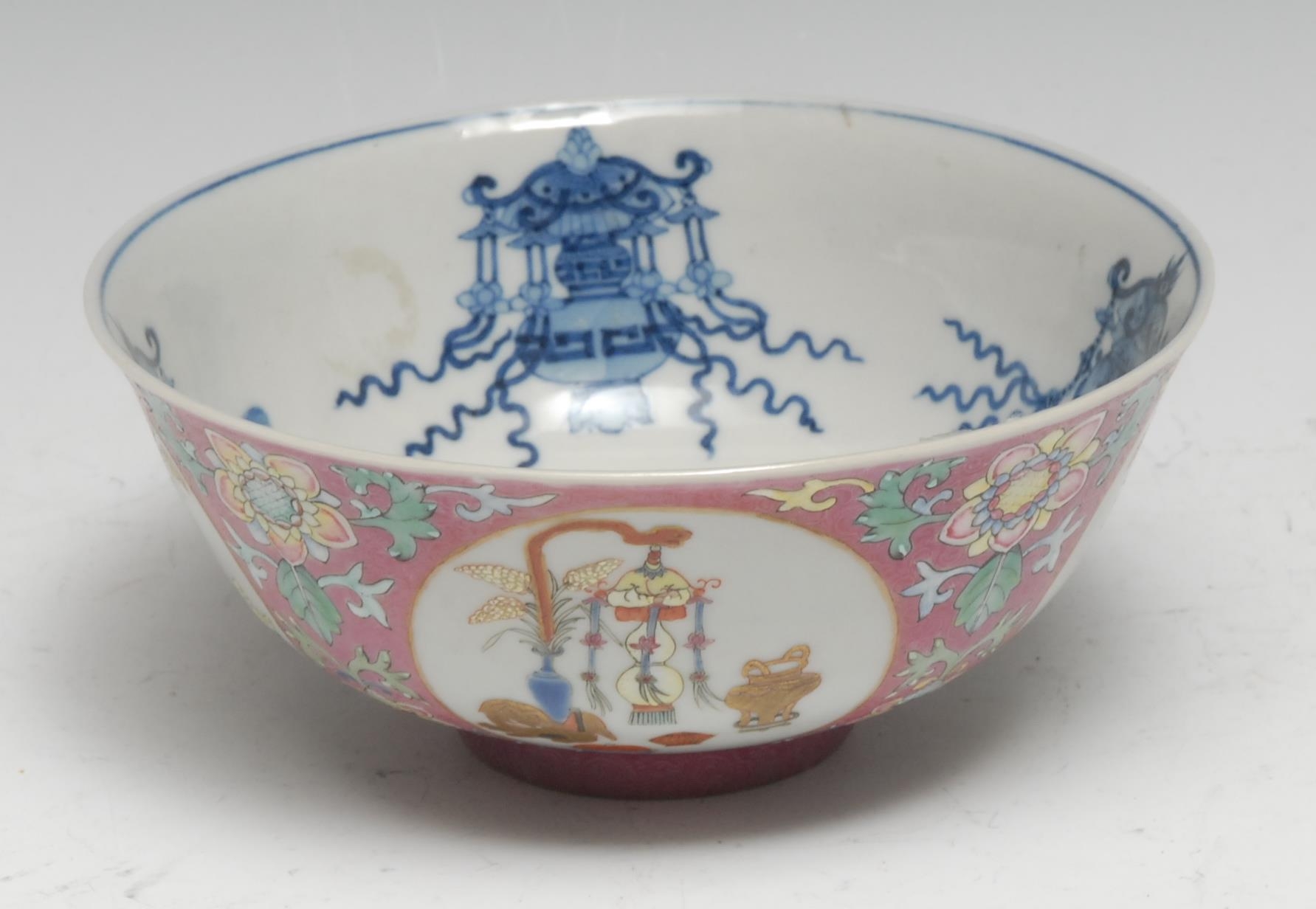 A Chinese Famille Rose porcelain flared circular bowl, painted with roundels of precious objects in polychrome enamels and picked-out in gilt, on a pink ground incised with leafy C-scrolls and painted in over-glaze enamels with flowering leafy foliage, the interior painted with further precious objects and foliage in underglaze blue, 14.5cm diam, square seal mark, Qianlong six-character seal mark in underglaze blue, 19th century
