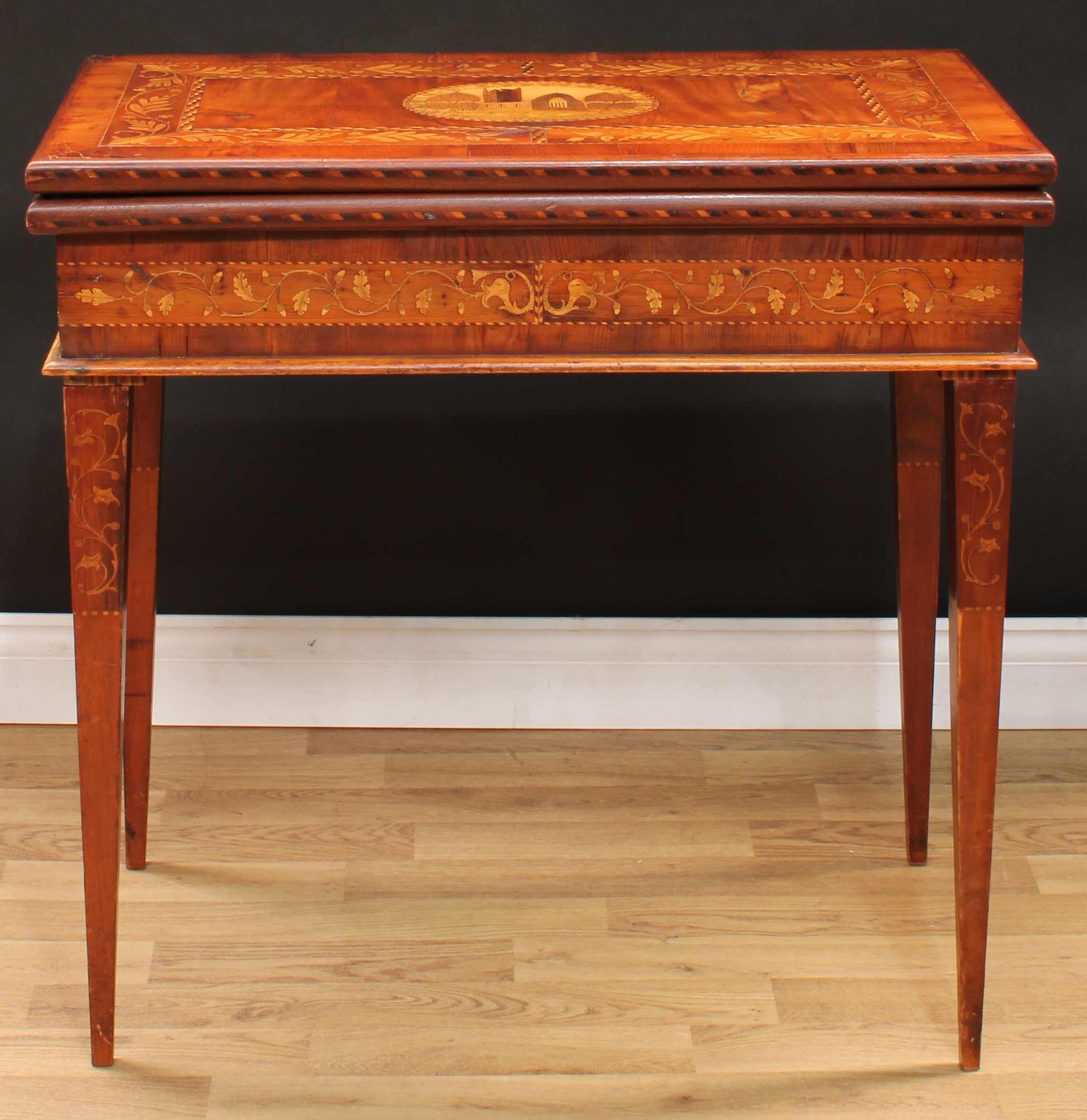 A 19th century Irish Killarney marquetry and yew room centre tea or silver table, hinged rectangular top inlaid with an oval reserve of Muckross Abbey, County Kerry, Ireland, bordered by trailing sprays of seamróg shamrocks, outlined throughout with barber-pole stringing and parquetry banding, enclosing further titled architectural landscapes of Innisfallen Abbey, Shooting The Rapids At The Old Weir Bridge, Ross Castle, Ross Cottage, Flesk Castle and Glena Cottage, above a deep frieze inlaid with oak leaves and acorns, tapered square legs, 80.5cm high, 81cm wide, 44cm deep, c.1850