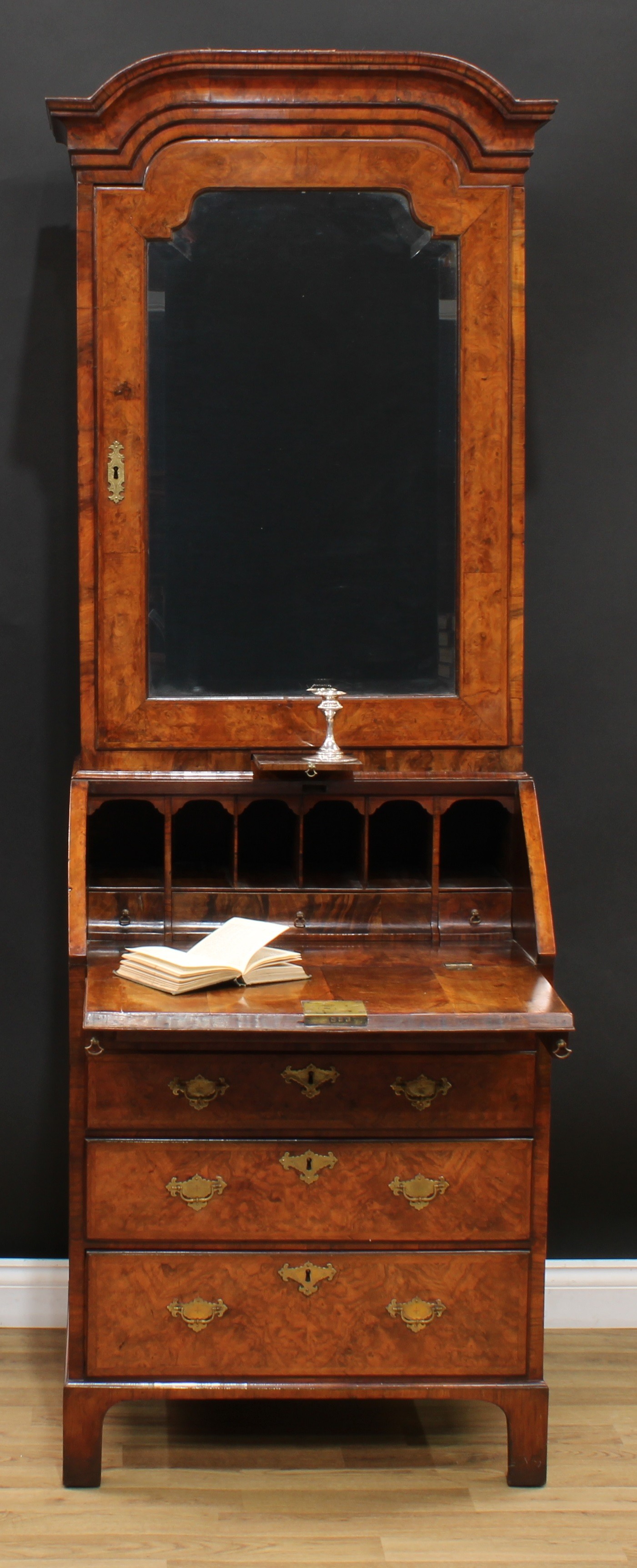 A George I walnut bureau book cabinet, domed cornice above a bevelled mirror door enclosing two adjustable shelves and a candle slide, the base with fall front enclosing three small drawers and pigeonholes, above four long graduated drawers, brass swing handles, brass foliate escutcheons, outlined throughout with herringbone stringing, bracket feet, 209cm high, 75.5cm wide, 51cm deep, c.1725