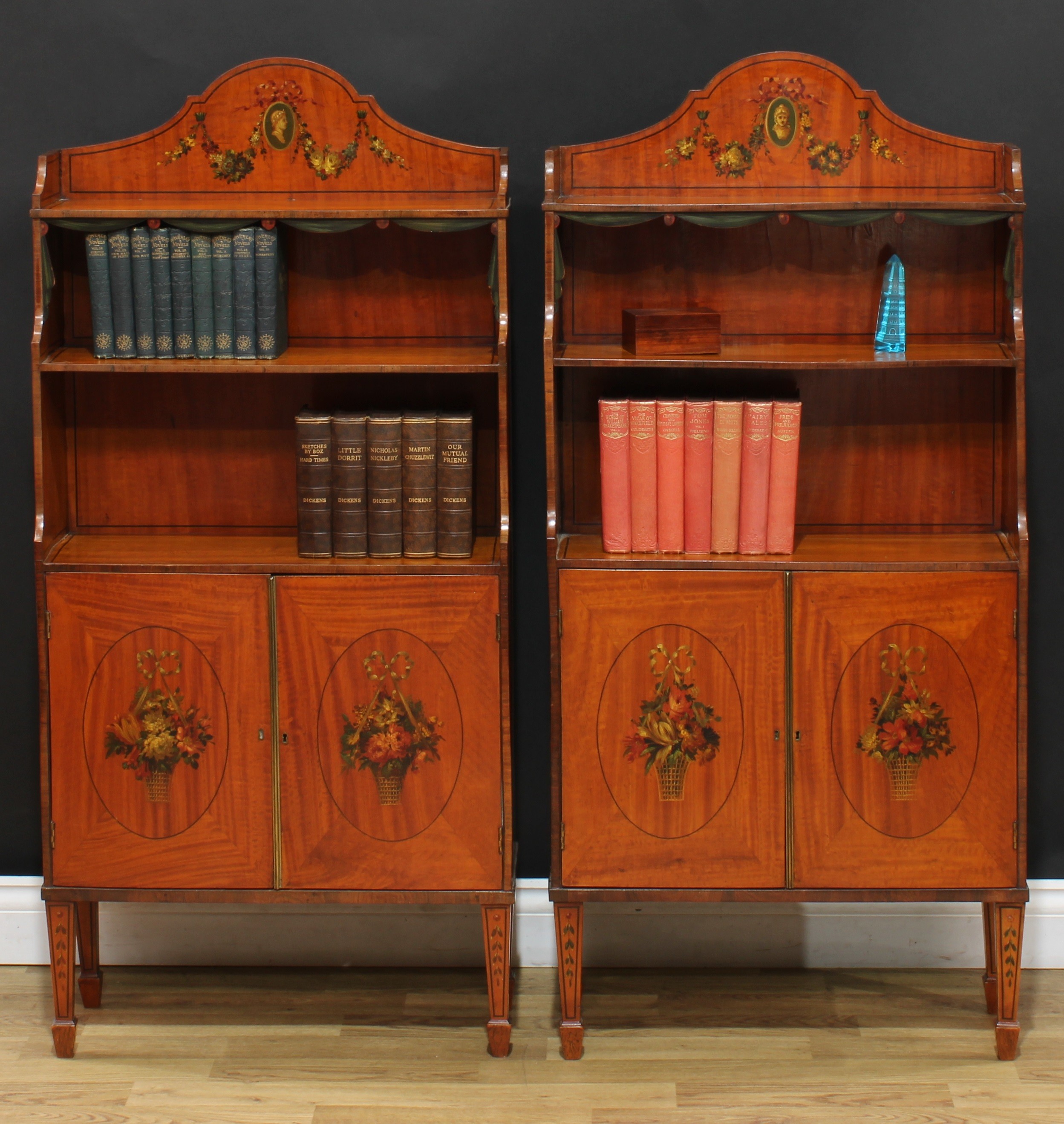 A pair of Sheraton Revival rosewood crossbanded painted satinwood waterfall bookcases, of small proportions, each with shaped arched gallery decorated with a ribbon-tied portrait and flowering swags, above three shelves, drapery swag friezes each base with a pair of cupboard doors painted with baskets of flowers, tapered square legs, spade feet, 125cm high, 59cm wide, 22cm deep, c.1890