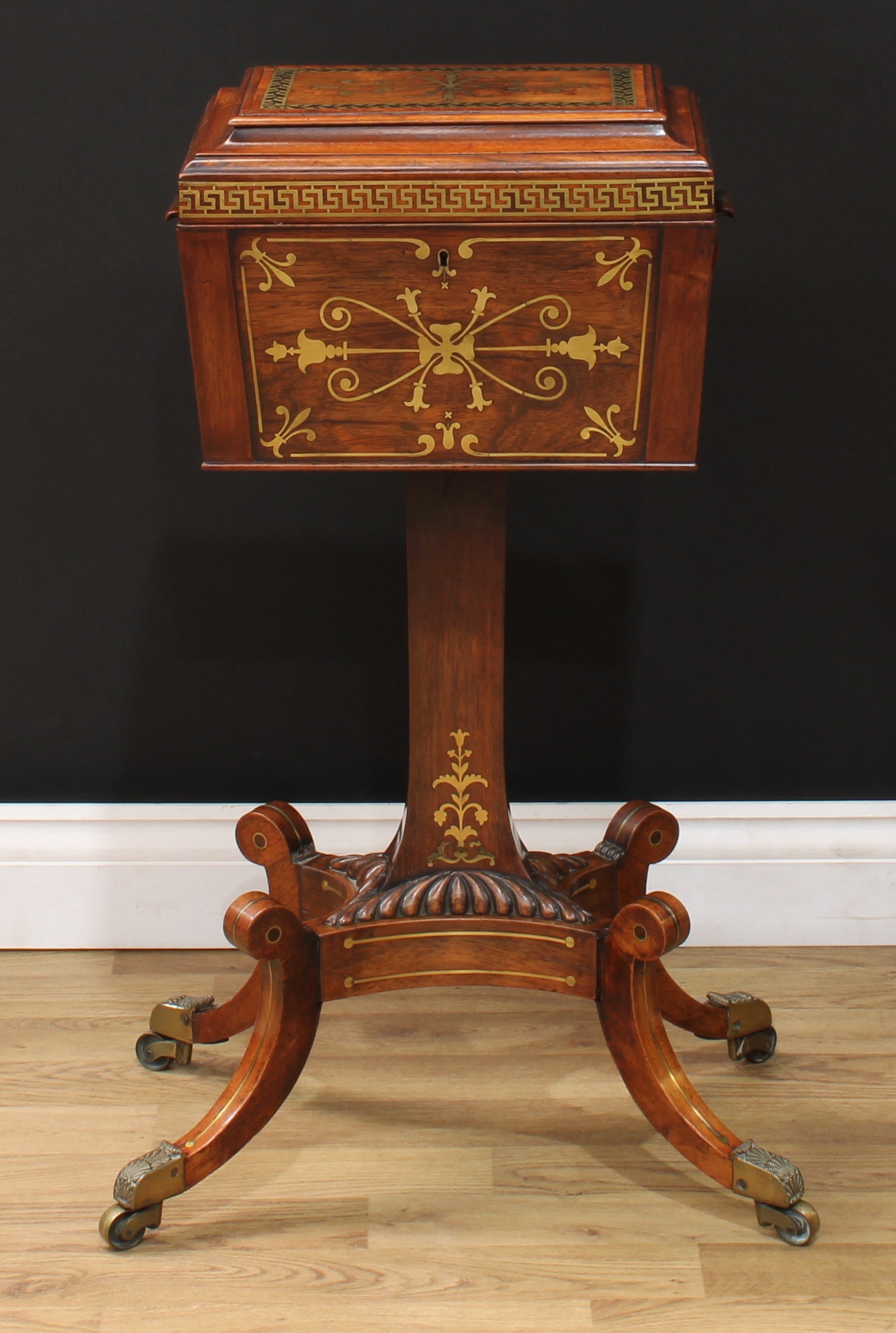 A Regency rosewood and brass marquetry teapoy, hinged sarcophagus caddy inlaid with stylised leaves and bell husks, oak leaves and acorns and a meandrous band, the cover with tooled and gilt leather lining and enclosing three hinged removable canisters and an etched glass mixing or sugar bowl, carry handles to sides, rectangular column, gadrooned socle, incurve rectangular base, hipped sabre legs, shell cast brass capped casters, 79.5cm high overall, the tea caddy 24.5cm high, 35.5cm wide excluding handles, 25cm deep, c.1820