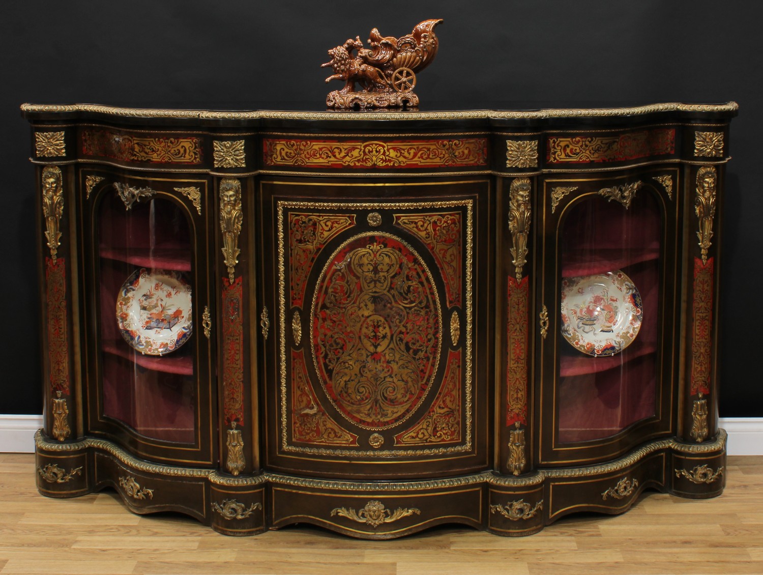 A substantial Victorian gilt metal mounted ebonised and 'Boulle' marquetry credenza, shaped top with egg-and-dart edge above a central panel door enclosing a shelf, flanked by serpentine glazed vitrine doors, each enclosing two further shelves, divided and flanked by cast and applied masks, inlaid throughout with scrolling leaves, plinth base, 114cm high, 207cm wide, 51cm deep, c.1880