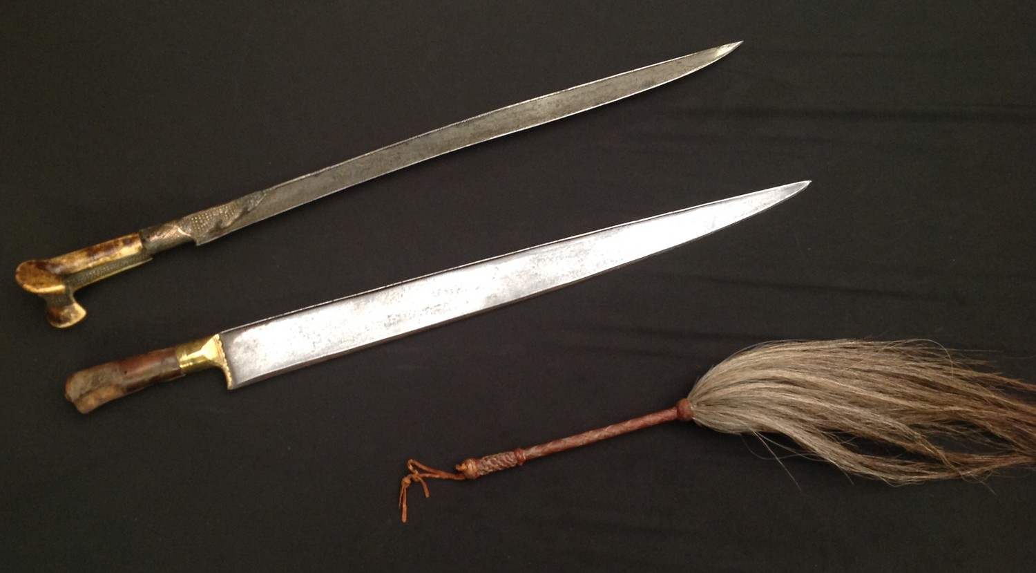 Pair of Indo-Persian Short Swords and a Horse hair fly swatter. First Sword has a ear style grip with single edged blade 530mm in length with gilt decoration. Horn grip. Some decoration missing from grip on one side. Overall length 675mm: the second sword has a single edged blade 542mm in length, wooden grip with some loss of wood to one side. Overall length 680mm. Neither swords has a scabbard; Fly swat is approx 560mm in overall length with leather bound grip, lanyard loop a/f (3)