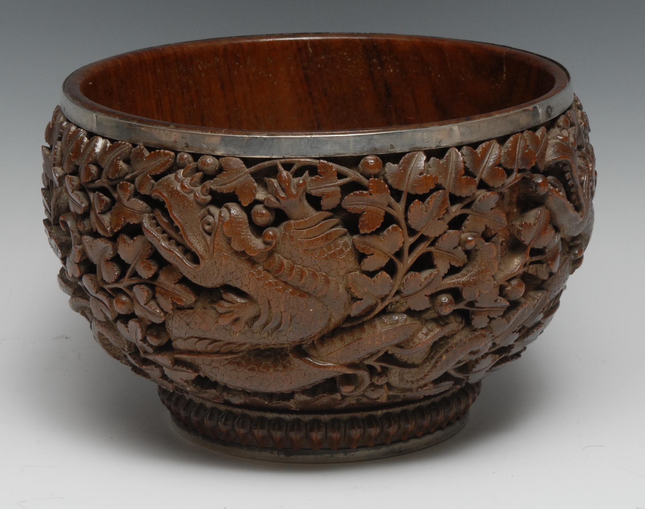 A Burmese silver coloured metal mounted hardwood bowl, boldly carved with dragons amongst dense fruiting branches, 23cm diam, c.1880