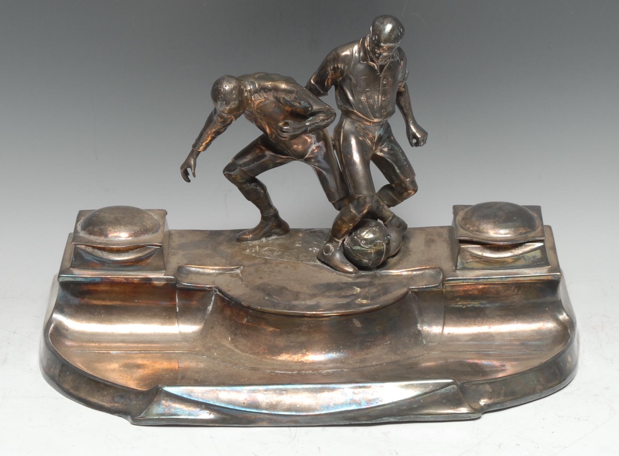 An Art Nouveau period silver plated inkstand, in the manner of WMF, surmounted by two football players contesting the ball, flanked by a pair of wells with hinged covers, 34cm wide, c.1910