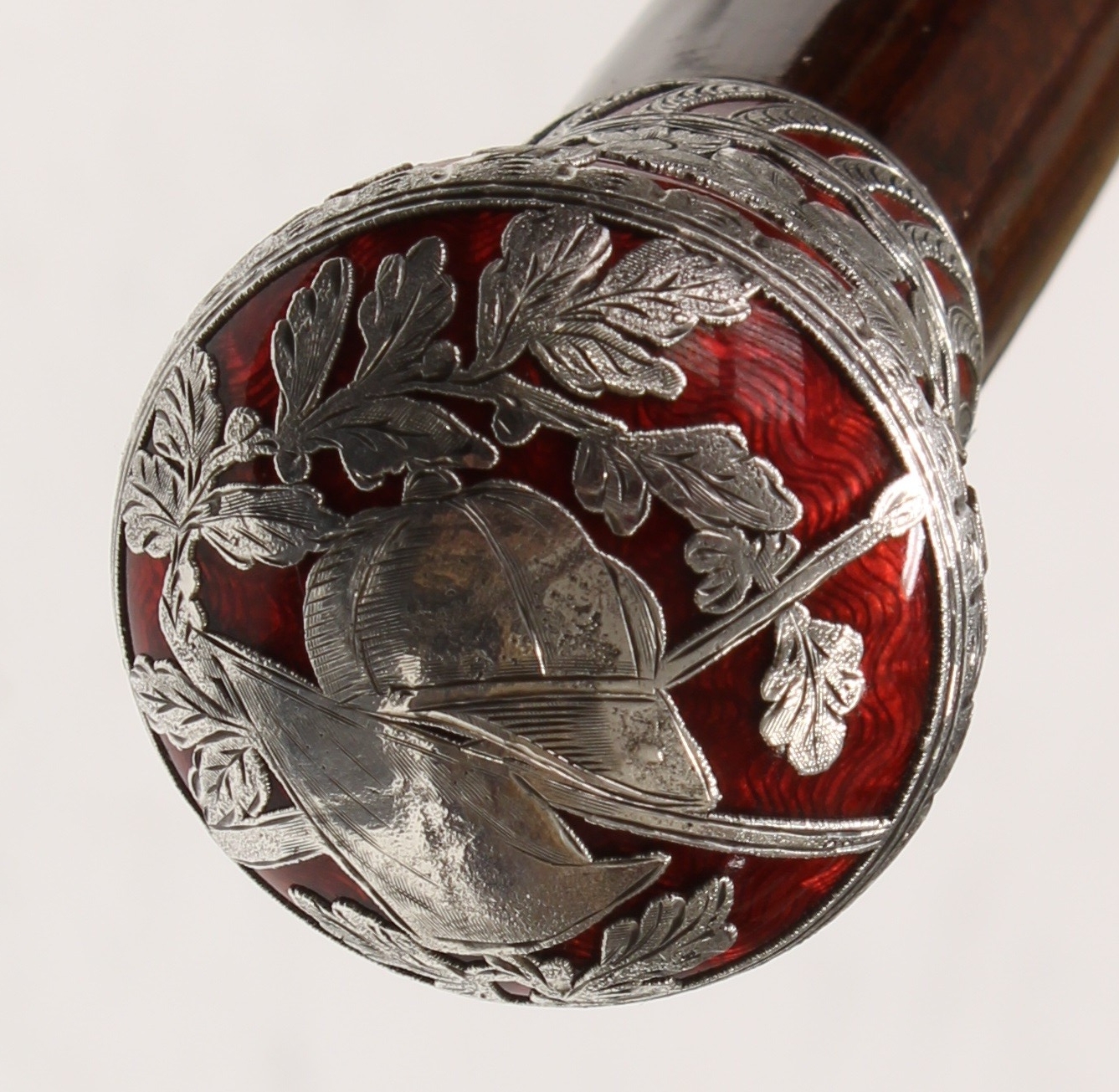 A French Belle Époque silver-mounted and guilloché enamel gentleman's walking stick, the globular pommel chased and pierced with a jockey's cap over a crossed horse riding crop and a racing flag, surmounted by acorn and oak leaves, above a frieze of flowering leafy foliage, the silver overlaid on a red enamel ground, tapering snakewood cane, horn walking tip, 95.5cm long, the handle c. 1900