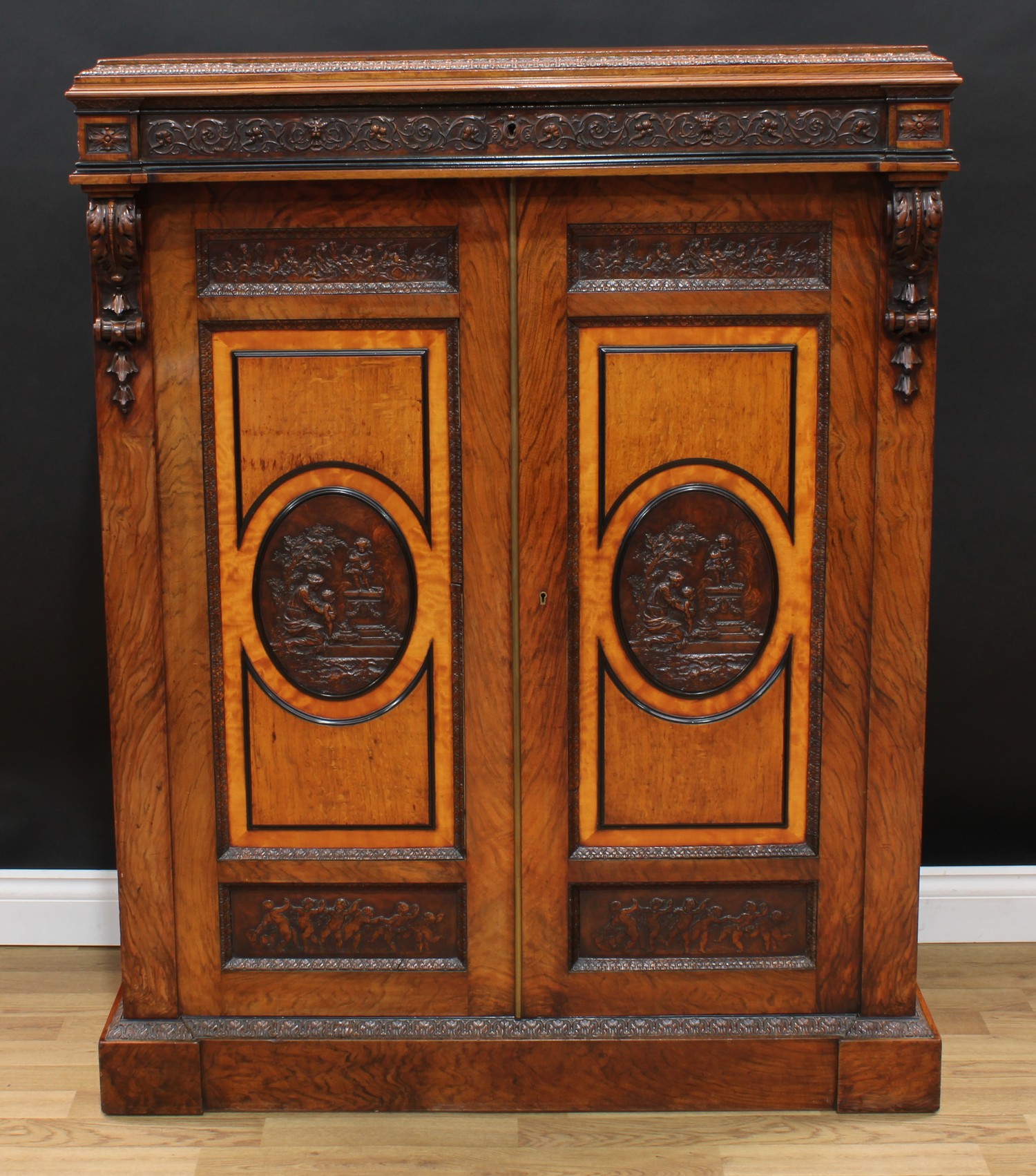 A Victorian walnut, oak and satinwood library cartonniere cabinet, in the manner of Holland & Sons, rectangular top with acanthus border above a frieze drawer in relief with Renaissance Revival foliage masks and leafy scrolls, above a pair of rectangular doors, with bas reliefs and centred by oval panels of classical subjects, ebony borders, enclosing a bookcase interior of filing shelves, flanked by acanthus carved scroll brackets, elaborate security hinges, the lock stamped Tucker & Reeves, VR patent and numbered 29142, plinth base, 139cm high, 107cm wide, 45cm deep, c.1870