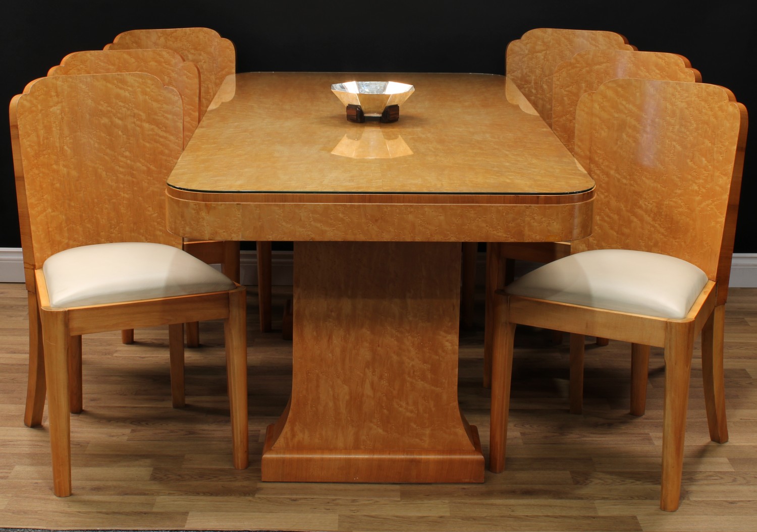 An Art Deco birdseye maple dining table, rounded rectangular quarter-veneered top, above a deep frieze, spreading rectangular supports, plinth feet, 76.5cm high, 182.5cm long, 91cm wide; a set of six conforming cloud-back dining side chairs, each with a curved back with reentrant ears, drop-in seat, tapered forelegs, 90cm high, 48cm wide, the seat 39cm wide and 39cm deep, possibly Harry and Lou Epstein, c.1930 (7)