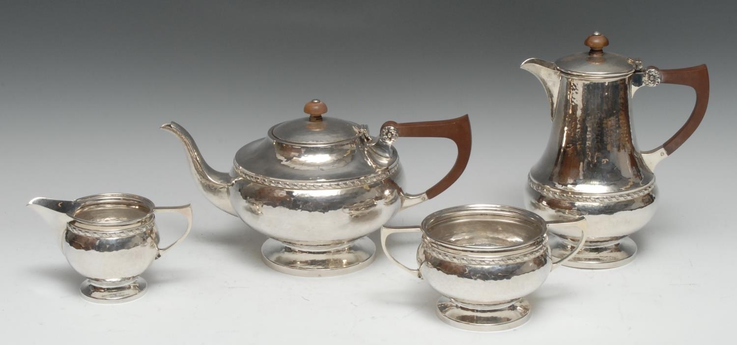 A E Jones - an Arts and Crafts silver four piece tea service, comprising teapot, hot water pot, milk jug and sugar basin, planished overall, ribbon girdles, angular scroll handles, bakelite fittings, the water pot 20.5cm high, Birmingham 1924, 41.25oz gross