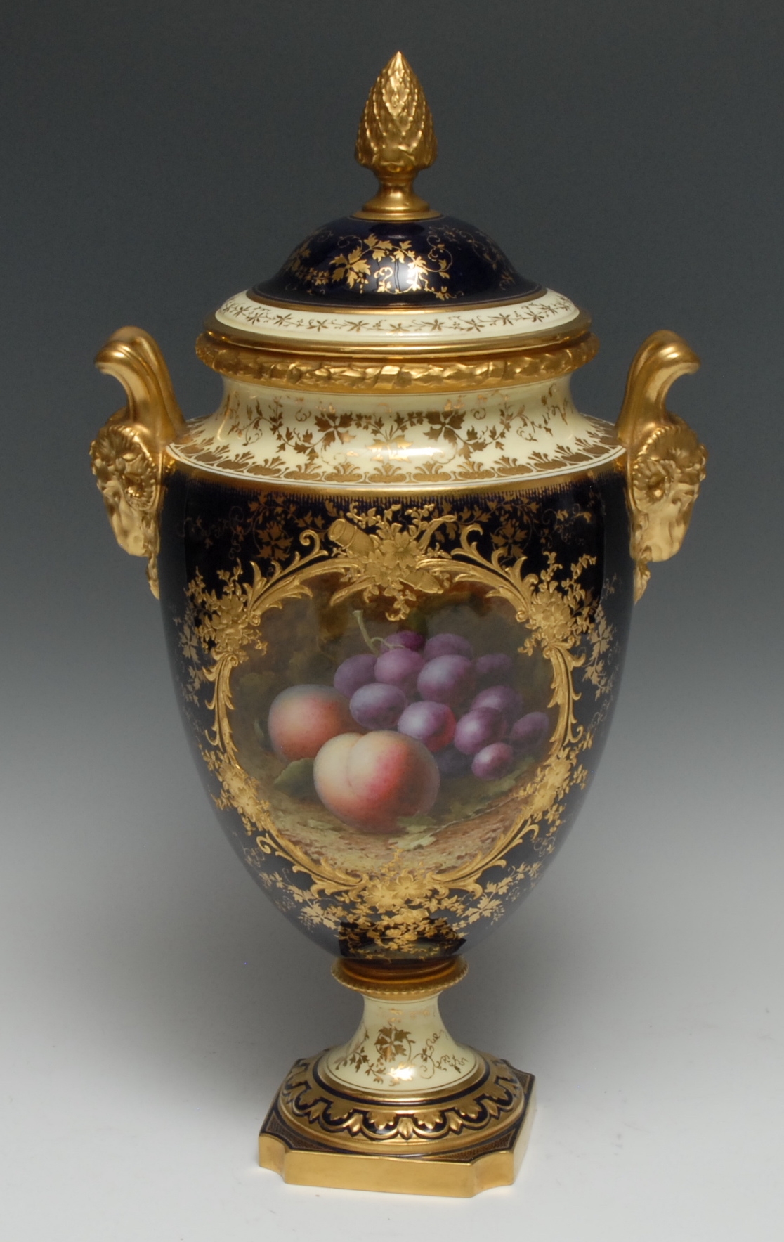 A Coalport two handled pedestal ovoid vase and cover, painted by Frederick Herbert Chivers, signed with peaches and grapes, withing a raised gilded cartouche, on a cobalt blue and lemon ground, gilt ram's mask handles, domed cover, bud finial, 41.5cm high, printed mark, retails stamp for Waring and Gillows
