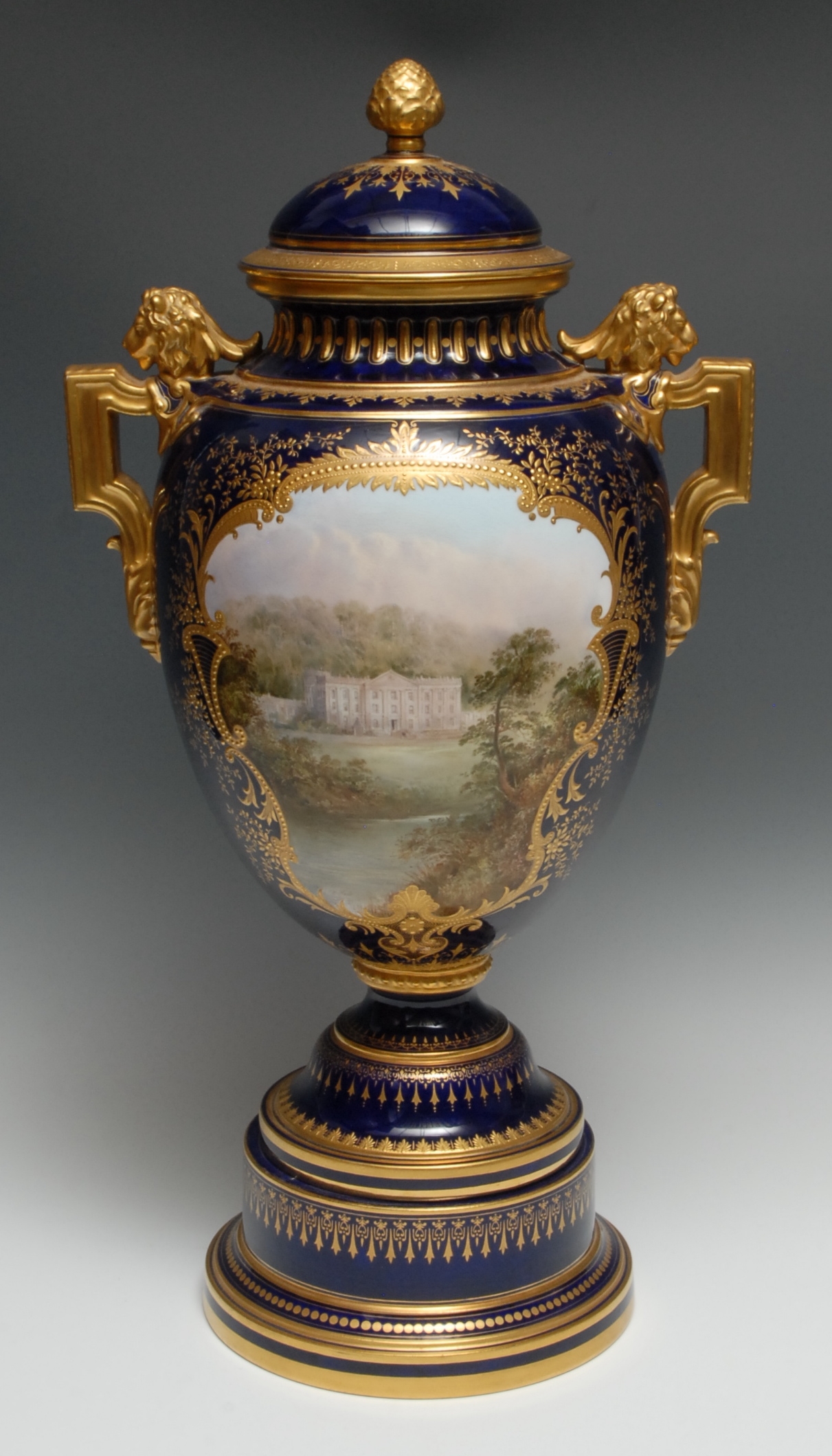A large and impressive Coalport vase and cover, painted by J H Plant, with Chatsworth House, with in jewelled gilt cartouche on a cobalt blue ground, lion mask and angular handles, domed cvoer, bud finial, pedestal base, 55cm high, pritned mark