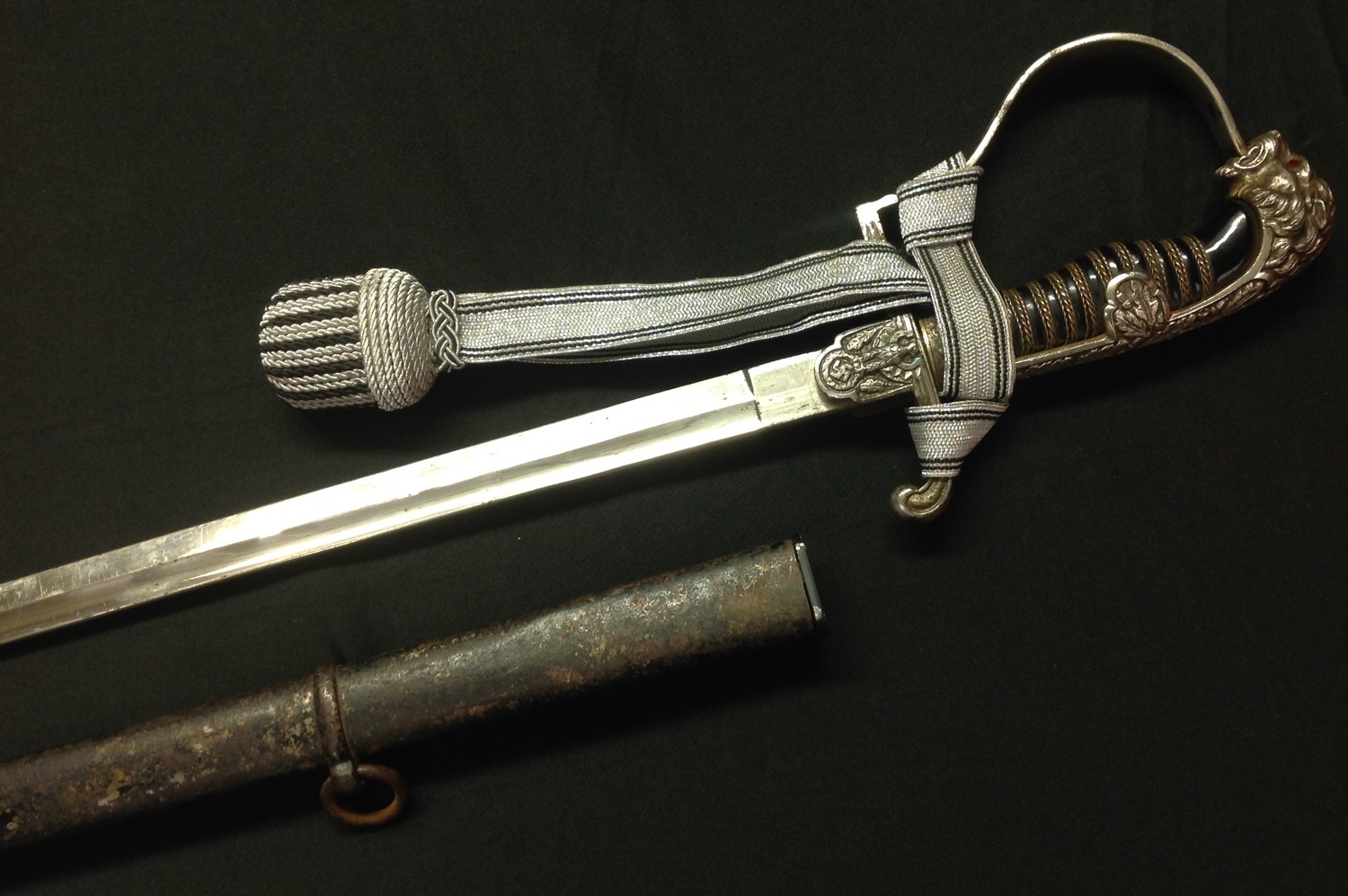 WW2 Third Reich Heer Officers Lion Head Sword with 860mm long fullered single edged blade, maker marked "Alcoso, Solingen". Wire bound black celluloid grip, Lion's eyes set with red glass. Silver finish to guard. Eagle to crossguard. Complete with scabbard wth remains of original black paint. Reproduction sword knot.
