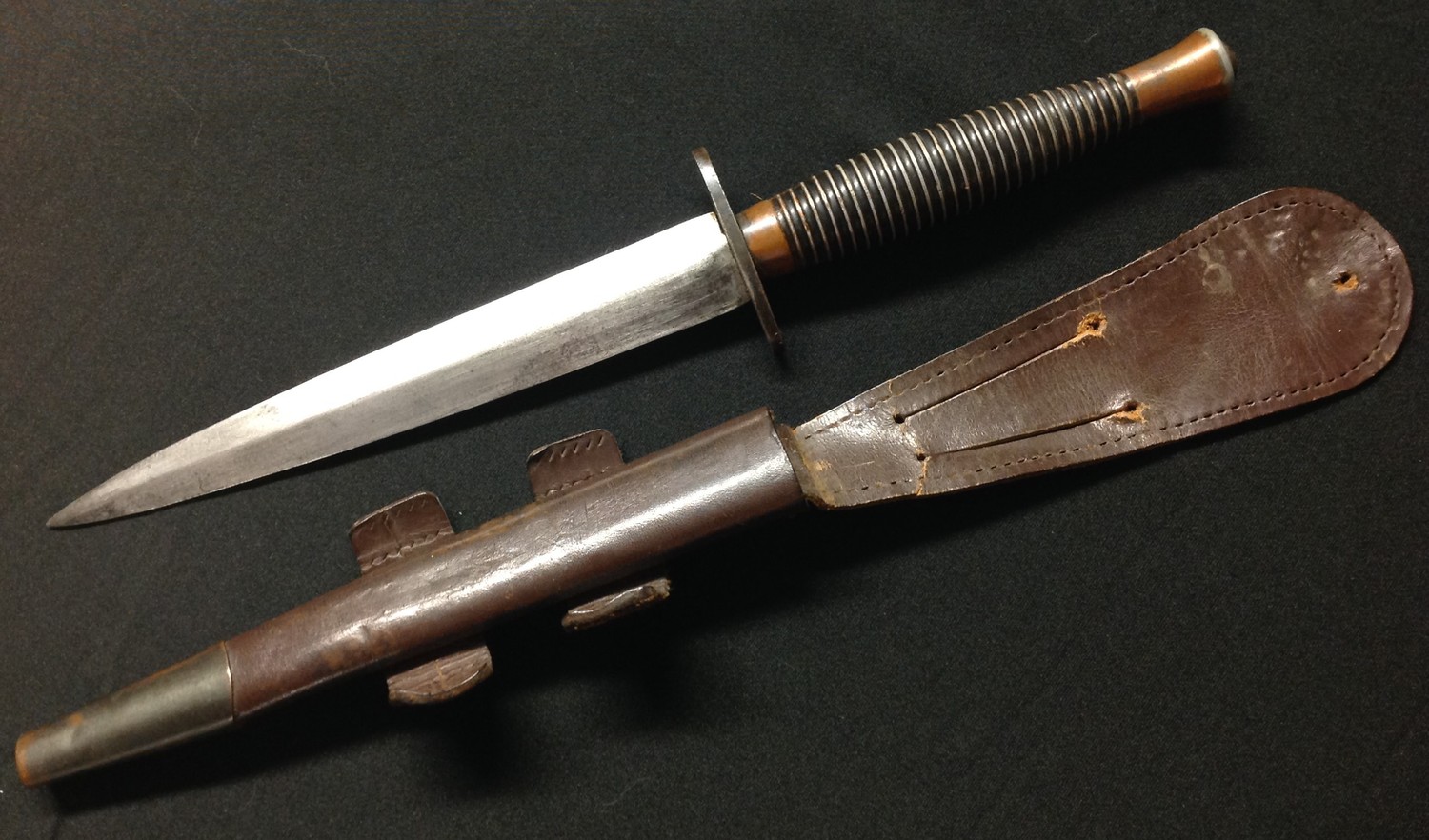 British Third Pattern Fairbairn Sykes Fighting Knife. 168mm long double edged blade with etched maker mark "Wilkinson Sword Ltd, Made in England". Crossguard unmarked. Cast alloy grip with copper undercoat finish showing, traces of the original black top coat finish remain to the grooves. Overall length 286mm. Complete with original scabbard with both leather tabs intact but missing elastic retaining loop. Blued steel chape.