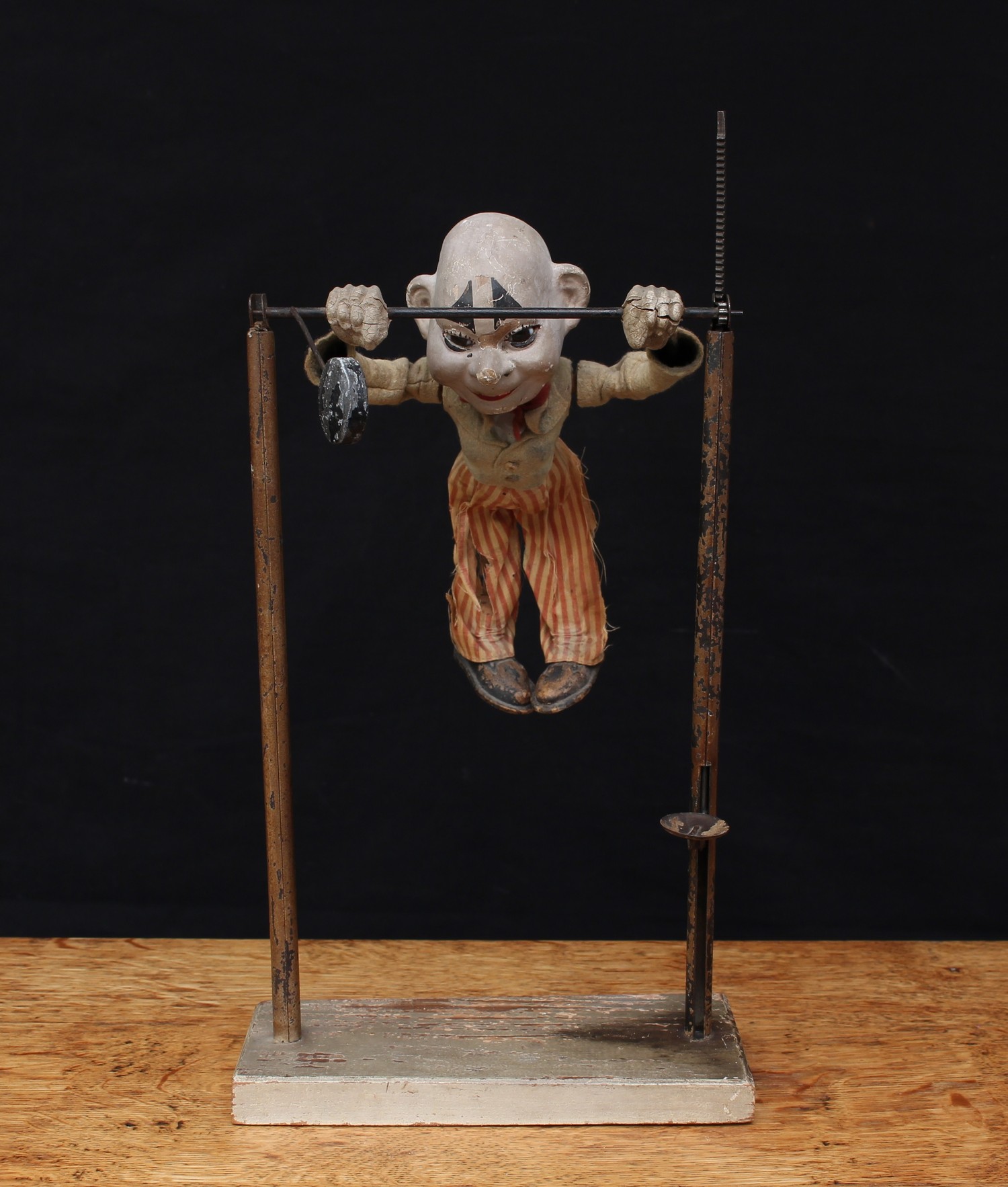 A late 19th/early 20th century novelty mechanical toy in the form of a clown or acrobat, painted papier-mache head inset with amber and black glass eyes, wearing a felt jacket with red bow tie and red and white striped trousers, mounted on a performance bar with a cog and rack driven mechanism, weight and thumb piece, rectangular wooden base, 39cm high