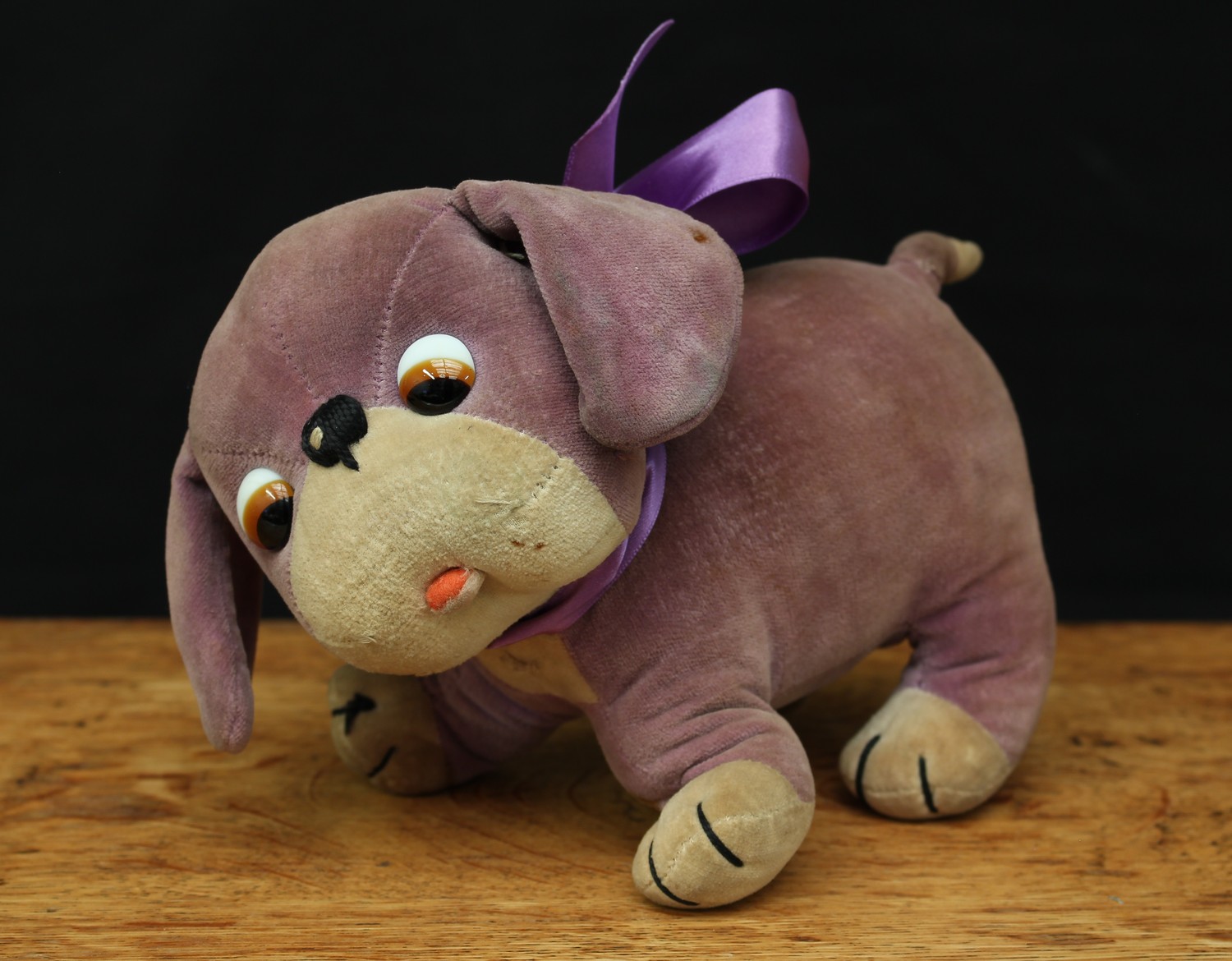 A J.K.Farnell 1920's novelty 'Dinkie' the Dog soft toy, mauve/purple and cream body, amber and black 'googlie' glass eyes, vertically black stitched nose, later purple neck ribbon, 18.5cm long, designed by Miss Chloe Preston