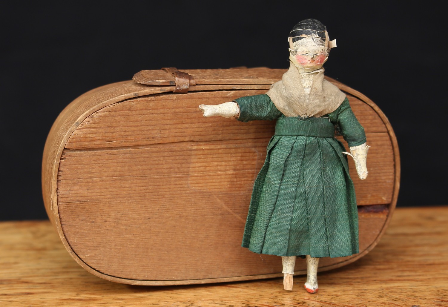 A mid-19th century miniature Grodnertal wooden doll, wooden head applied with gesso, painted features including rouge cheeks, wooden peg type body with gesso applied arms and legs, wearing a green silk dress, 9cm high, c.1860, stored in a small oval shaped bentwood tine box with cover